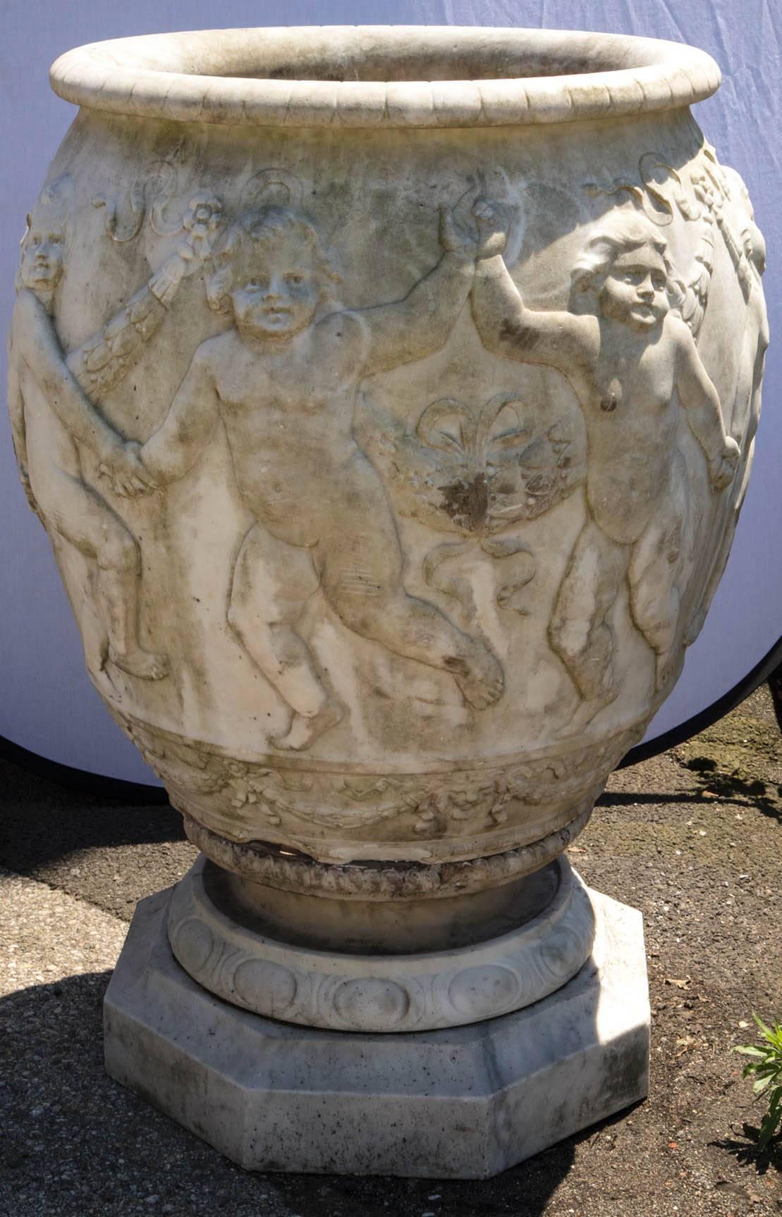 A pair of monumental antique marble urns. Each urn is carved with raised impressive cherubs depicting dancing. Eight cherubs on each urn, draped with ribbon/tassels. The carving is done very decoratively and shows great detail. The bases are octagon