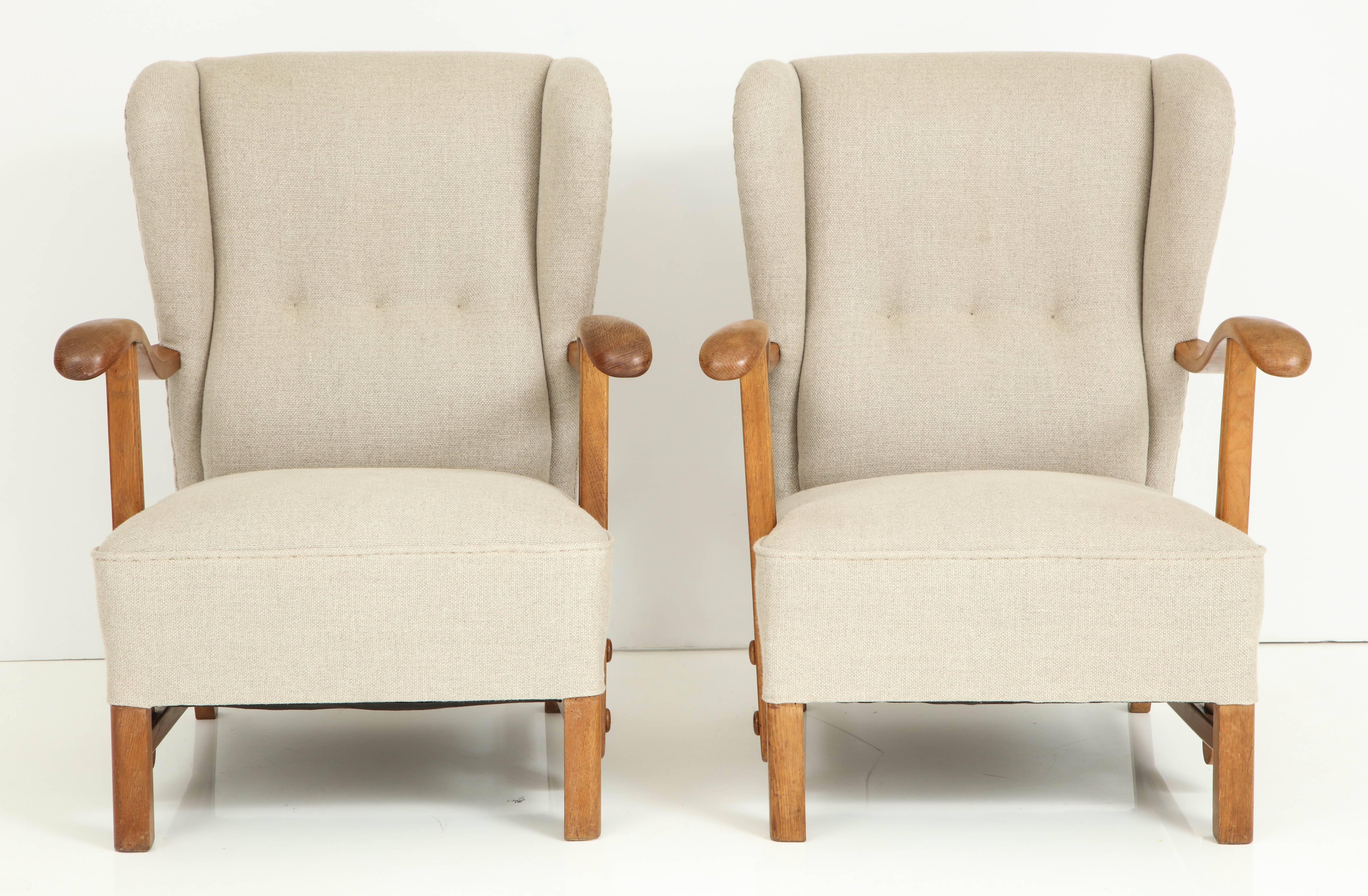 A pair of Danish oak and linen upholster open armchairs, circa 1940s, each with a tufted wing back, curved armrests raised on straight supports with dowels, and square legs joined by a stretcher.