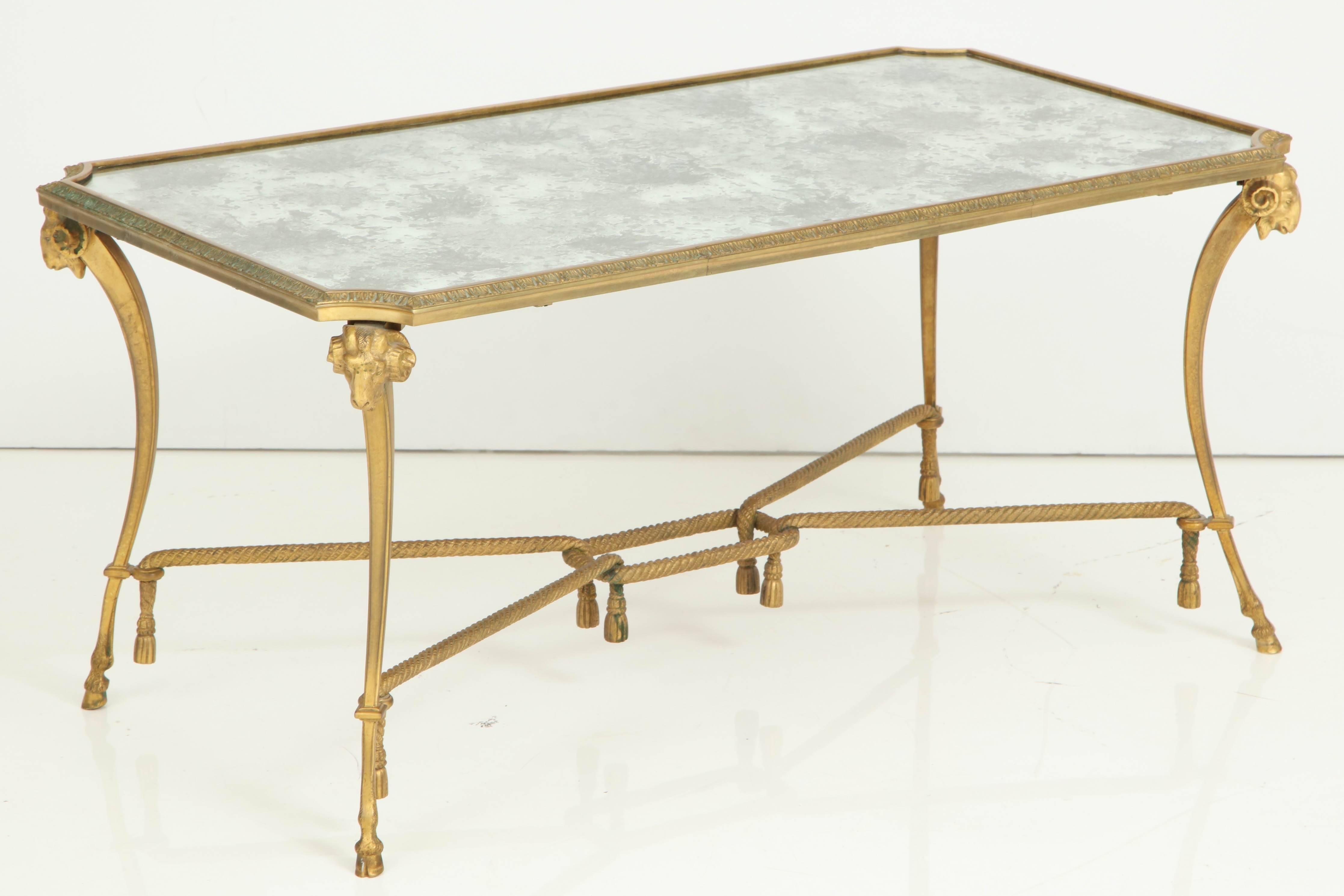 Empire style 1940s Baguès gilt bronze coffee table having distressed mirrored top, hoofed feet, scrolled supports with ram's head caps and tassel accents all joined by an h-stretcher.
         