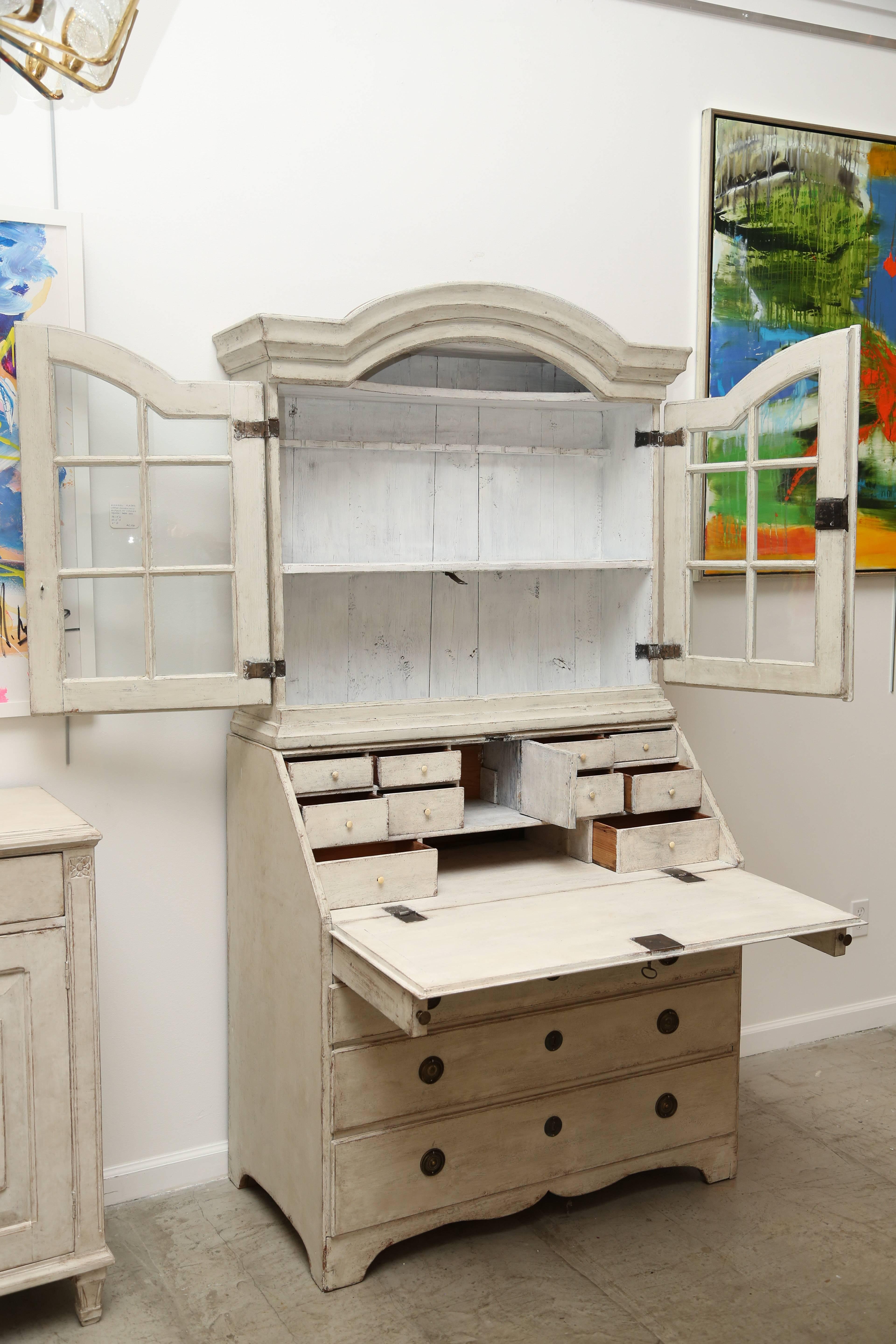 Antique Swedish Baroque Secretary with glass doors, painted Swedish distressed white/cream color. Upper section has curved arched doors with original glass panels, two shelves for books or display, a spoon rack and shallow top curved shelf for small