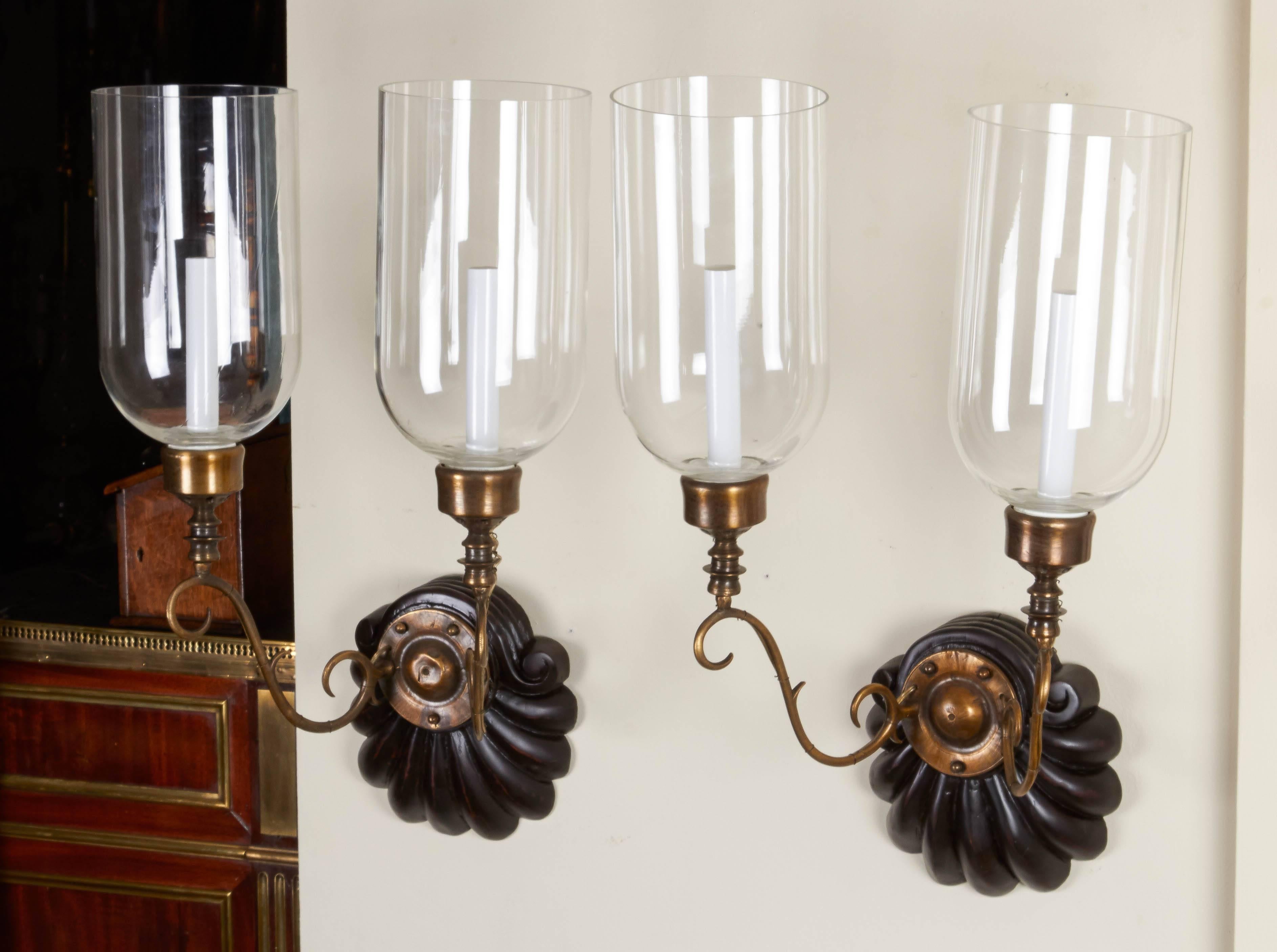 A pair of ebonized Anglo-Indian two-light sconces, shell form backplate issuing bronze out-scrolled candle-arms supporting a single Edison socket with hurricane shade, circa 1890.
Provenance- These sconces were purchased by Albert Nestle in India,