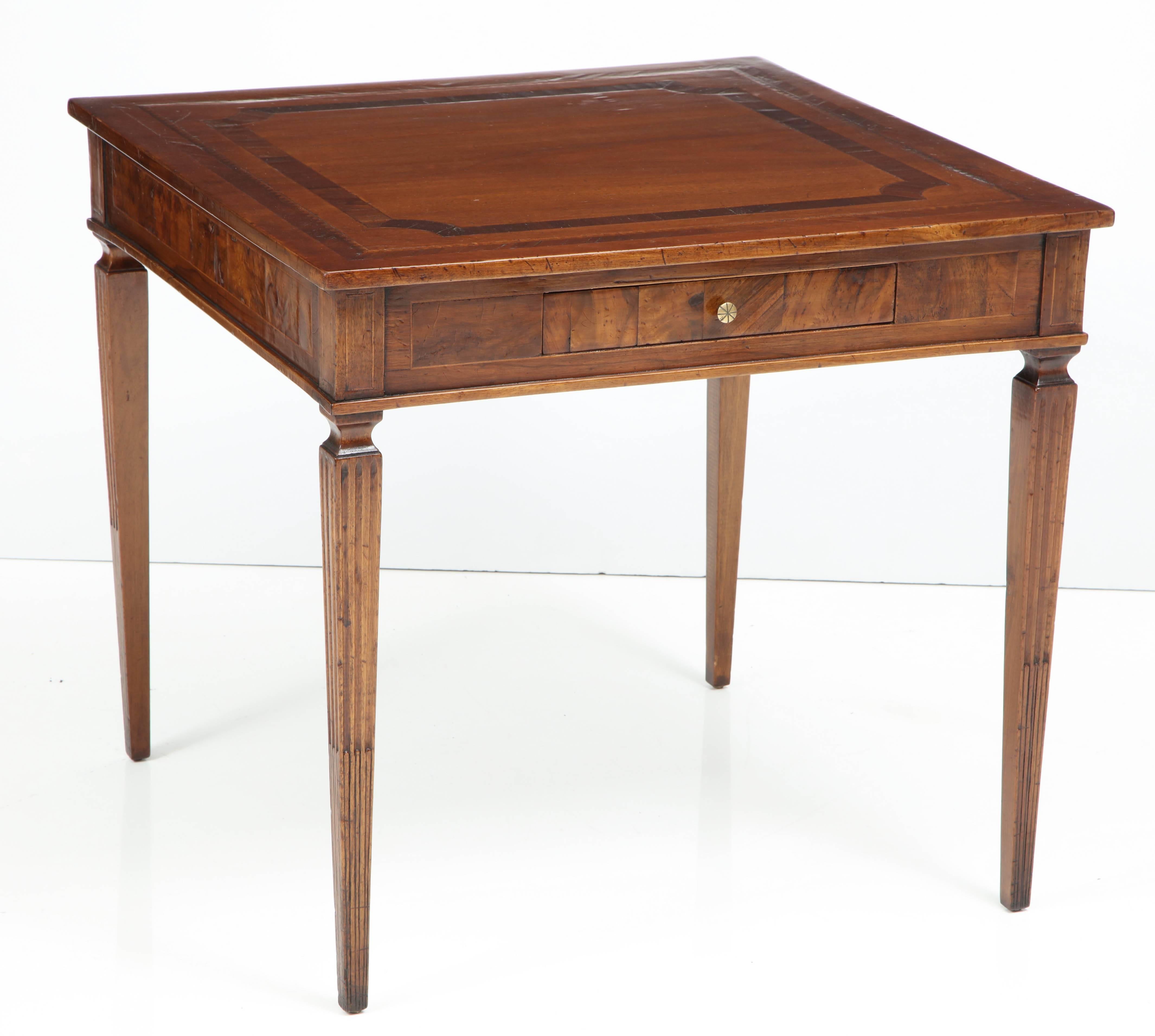 A walnut card table with elegant, Classic detail, including a beautifully crafted inlaid design on the top, a burl wood apron, and gracious tapered and fluted legs. The piece has one small drawer with a simple brass pull. It is finished on all sides