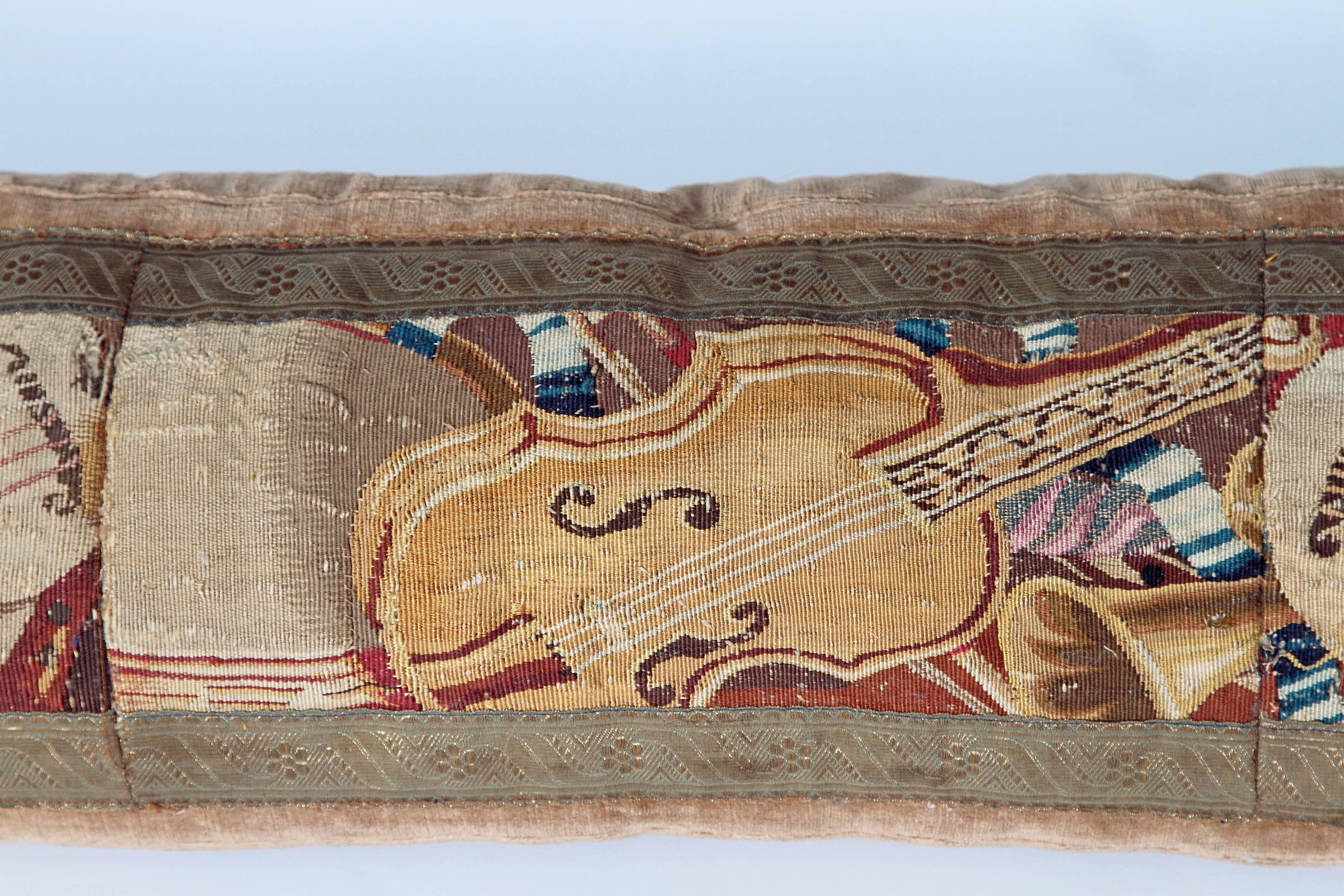A long beige velvet pillow with tapestry showing musical instruments in beiges and browns.