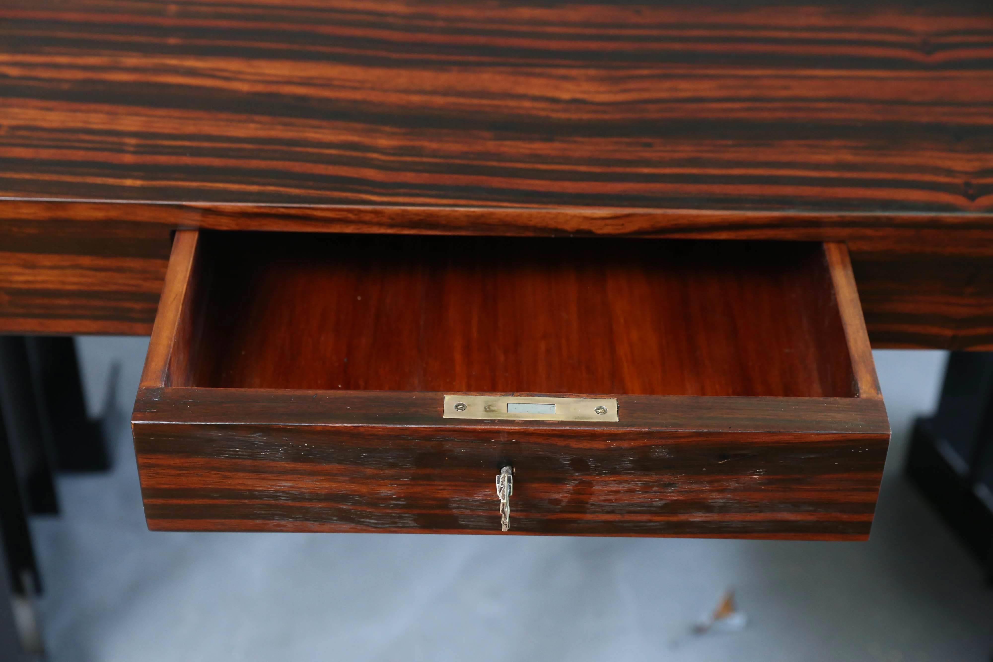 Das ungewöhnliche Design des Schreibtisches macht ihn zu einem einzigartigen Stück. Der Tisch ist aus Palisanderholz gefertigt und hat zwei Beine aus Metallplatten. In der Mitte der Tischfront befindet sich eine kleine Schublade. Die Tischplatte hat