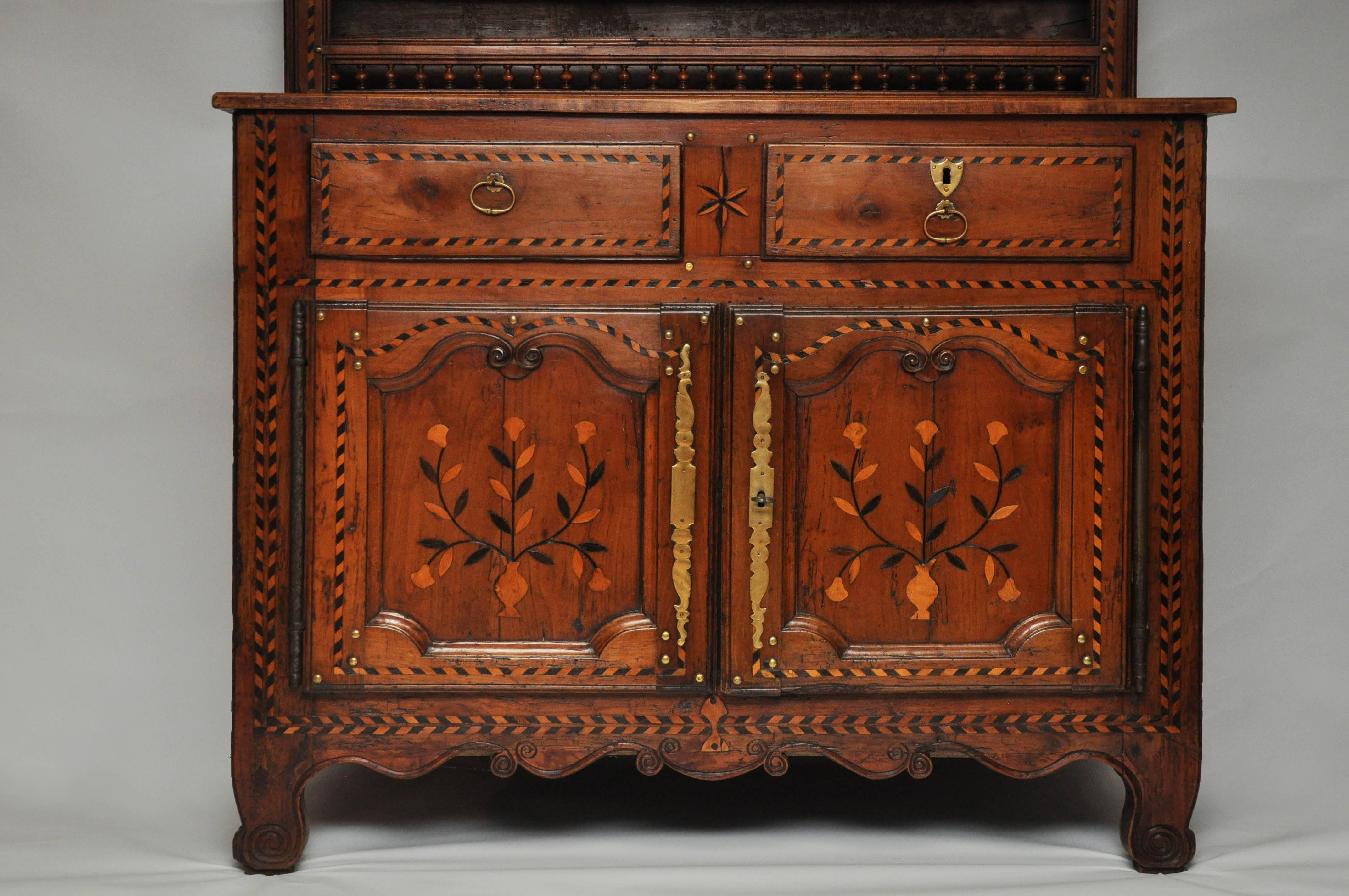 Hand-carved walnut chateau cupboard with a linear crown above a four shelf plate rail, each shelf having an elaborately carved spindle front support. The bottom portion consists of two drawers above a two door commode-all with intricate inlays of