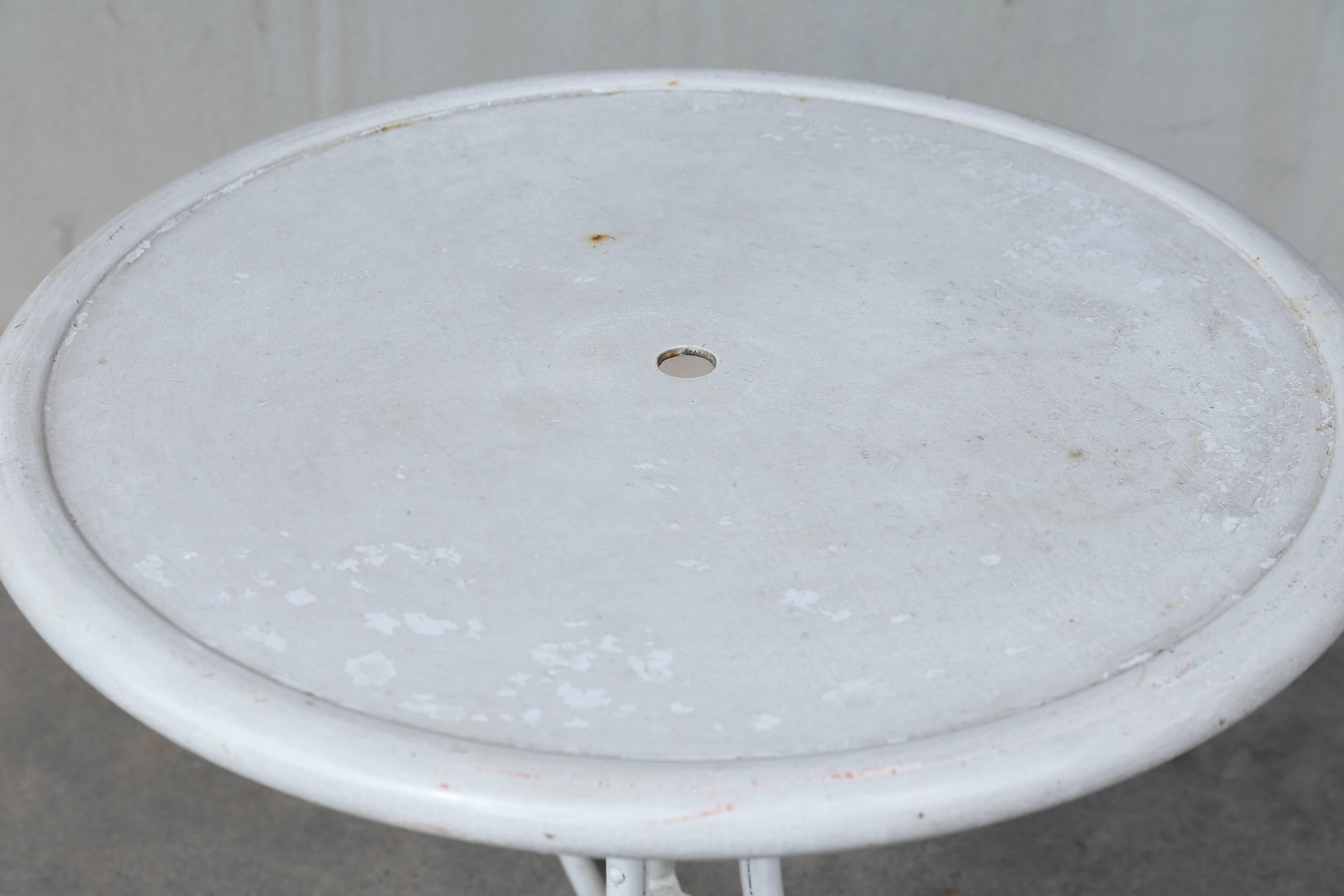 This is an early 20th century metal outdoor bistro table sourced in France. The table will accommodate an umbrella and features three curved legs. The top has a rounded edge and has been painted white. Some chips and scratches in the finish, and