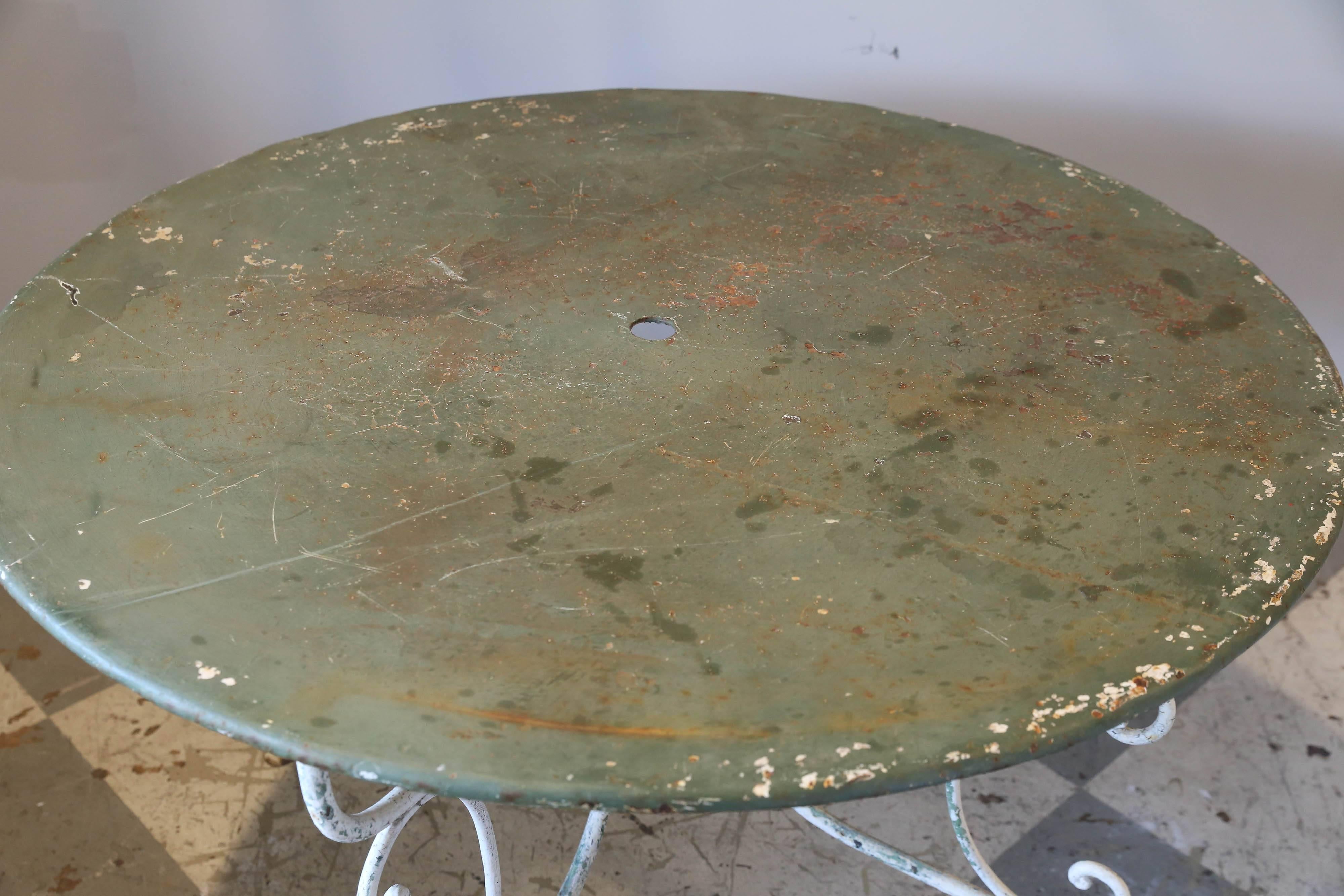 This is a rustic painted metal garden table we sourced in northern France. The base is painted an off-white color with some light green as well as the rust color of the metal underneath. The top is painted in a medium green. The centre of the top