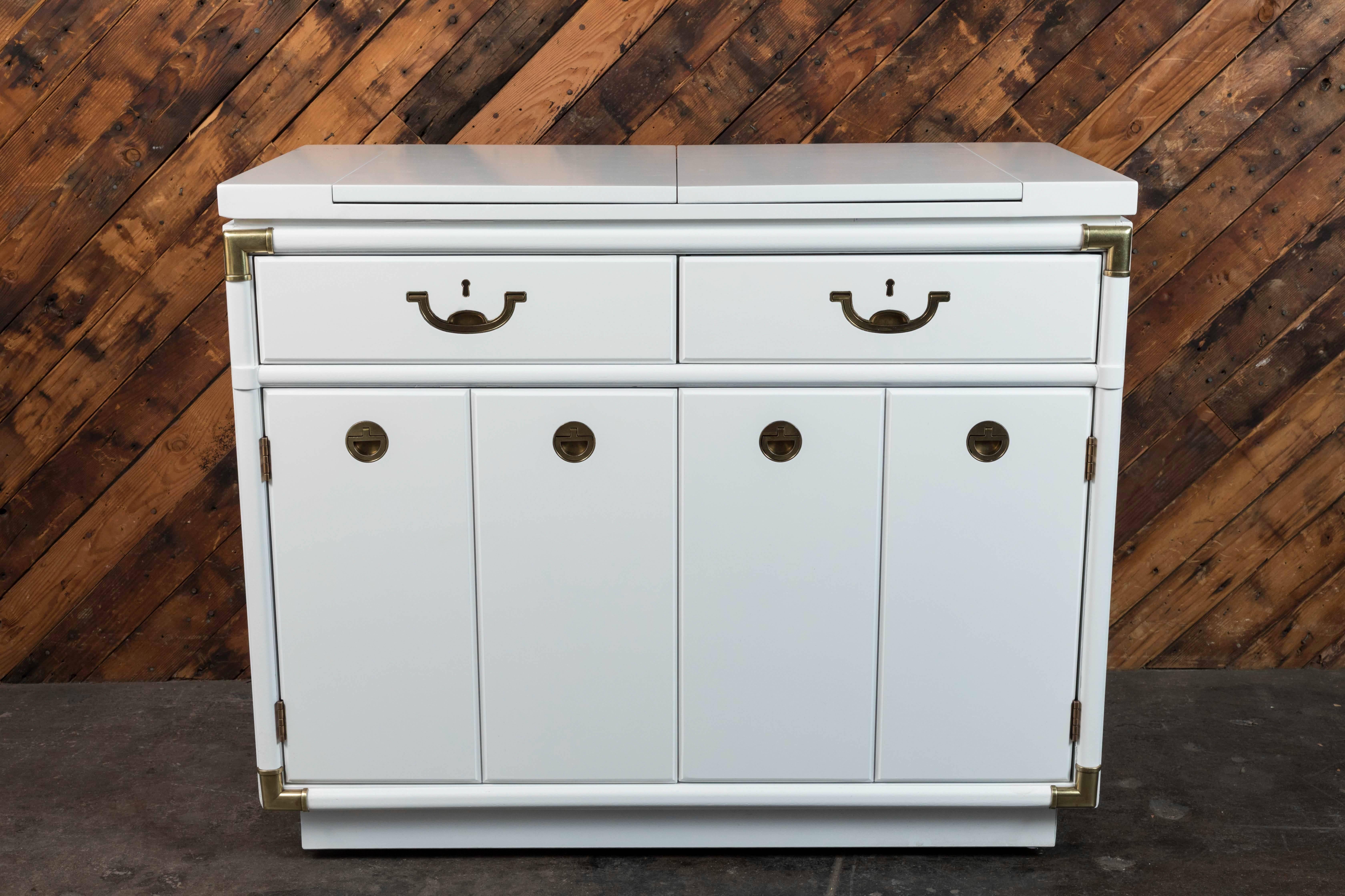 Gorgeous midcentury Campaign style rolling bar cabinet from the Drexel Accolade collection. Has two leaves that flip open. Plenty of storage. Note key holes are simply decorative. Has just been lacquer painted white and brass has been polished. When