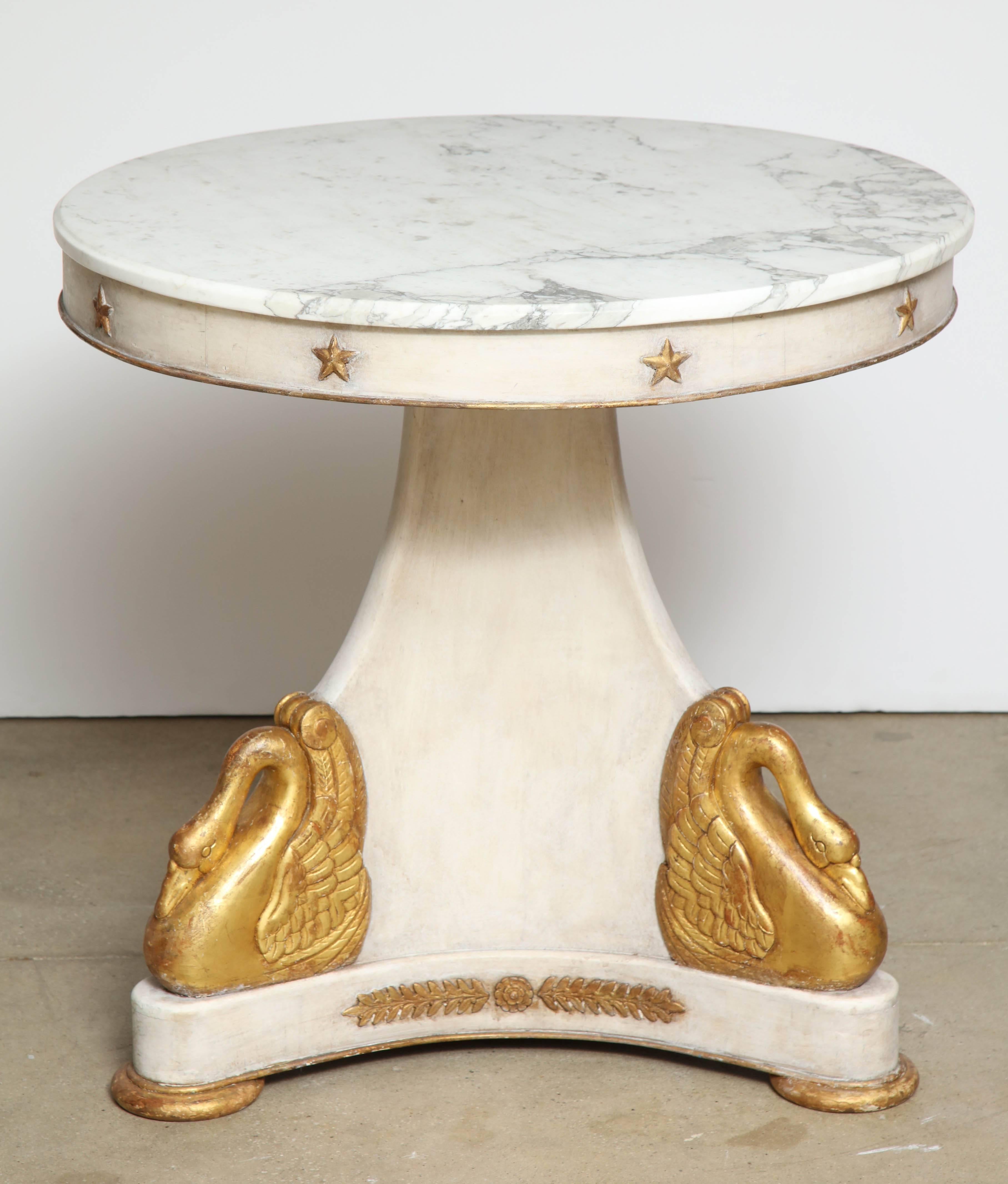 A most unique and unusual round table. The center pedestal base is painted in a grayish white featuring three stunning swans in a gold leaf finish, a detailed laurel leaf design, and three gilt round feet. The apron is painted and accented with gilt