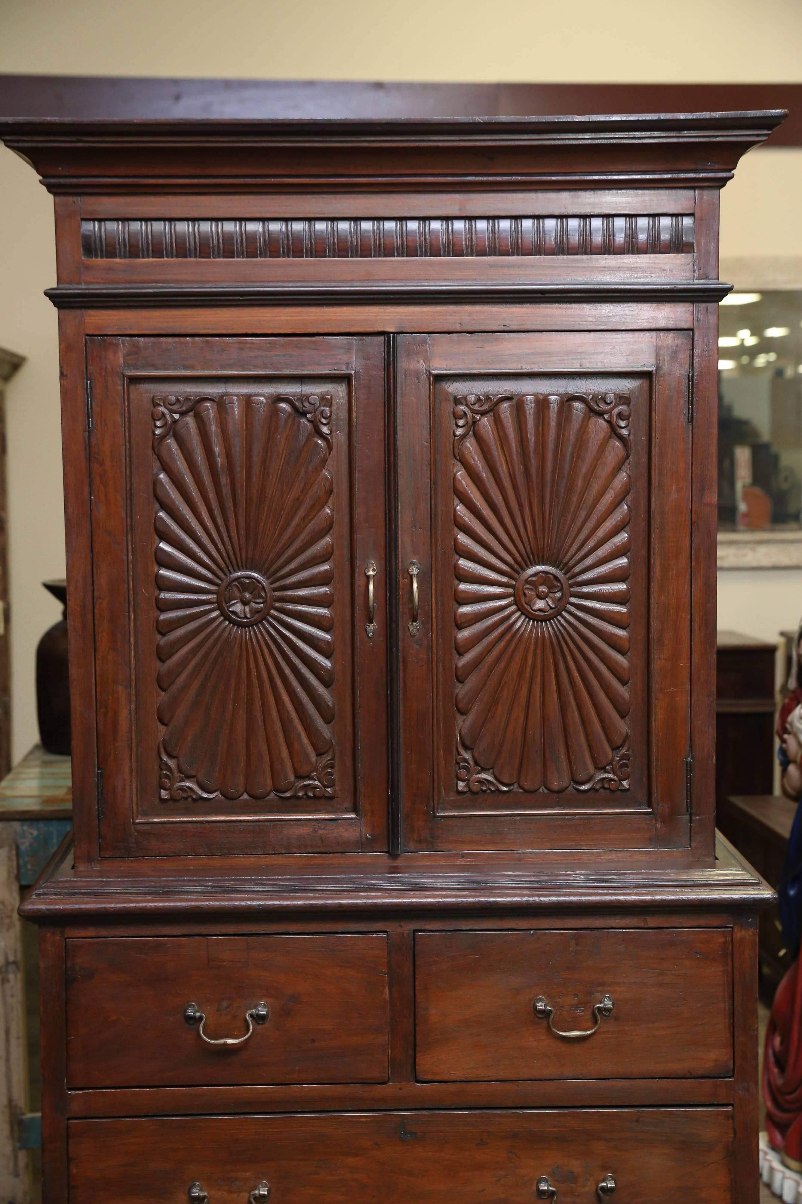 This fine teak wood cabinet has shelves on the top with two inside drawers and two small and two large drawers at the bottom. The doors at the top are carved with sunburst motif. It stands on sliding bracket
support. Comes from a British settler's