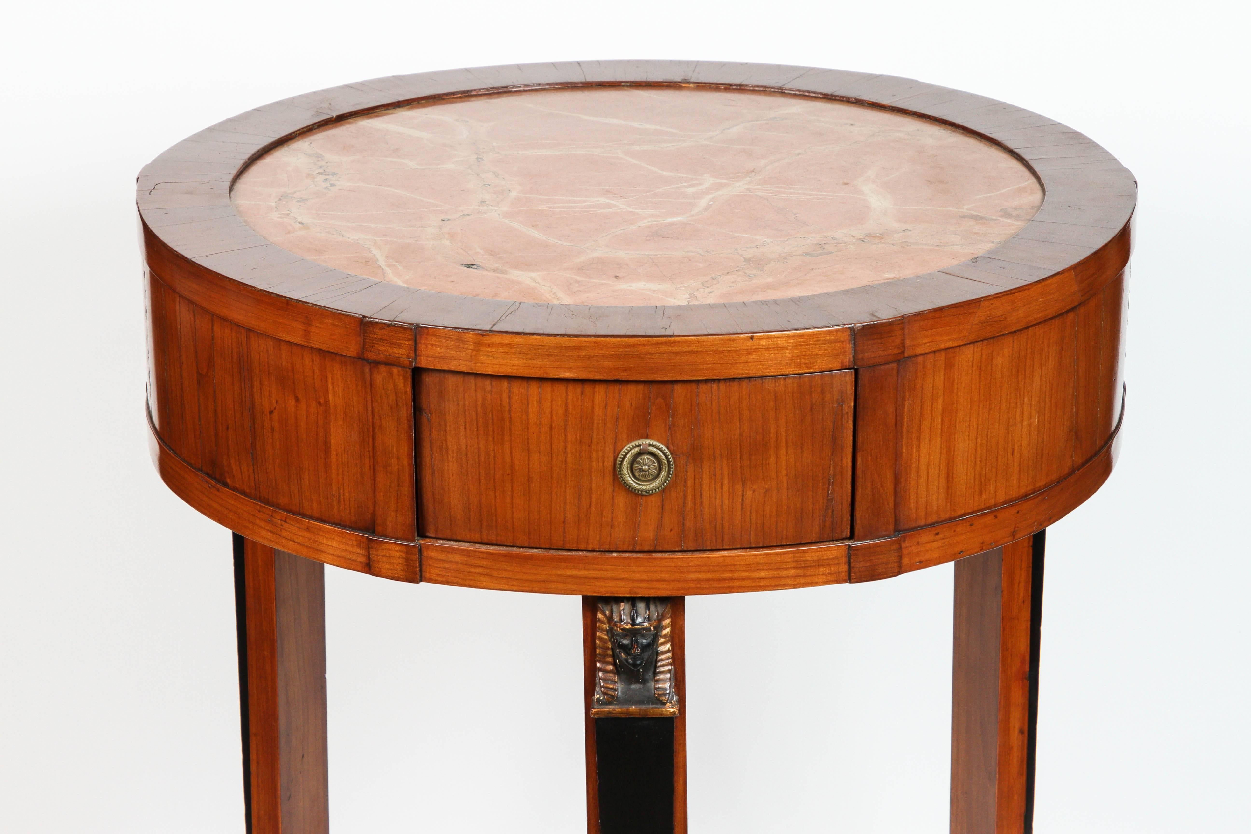 A beautiful Italian walnut neoclassical round table with inset marble top, drawer, and brass hardware. The table is raised by paws joined by a triangular stretcher with concave sides. Each leg has a carved pharaoh-figure.