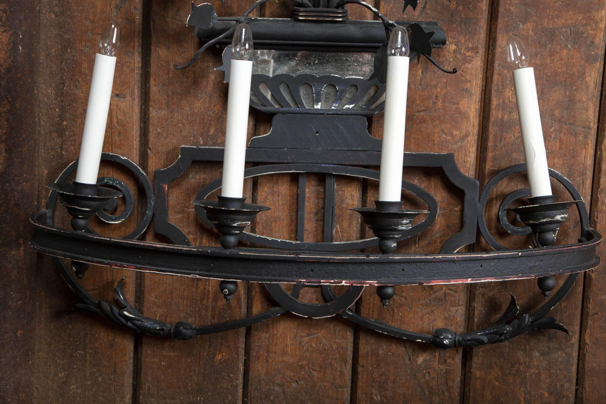 Large stylish black wrought iron wall sconce. It is electrified with seven candles. It would also be great with real candles. There is a decorative urn with flowers below the round distressed mirror. The sconce is topped with scrolls. A nice