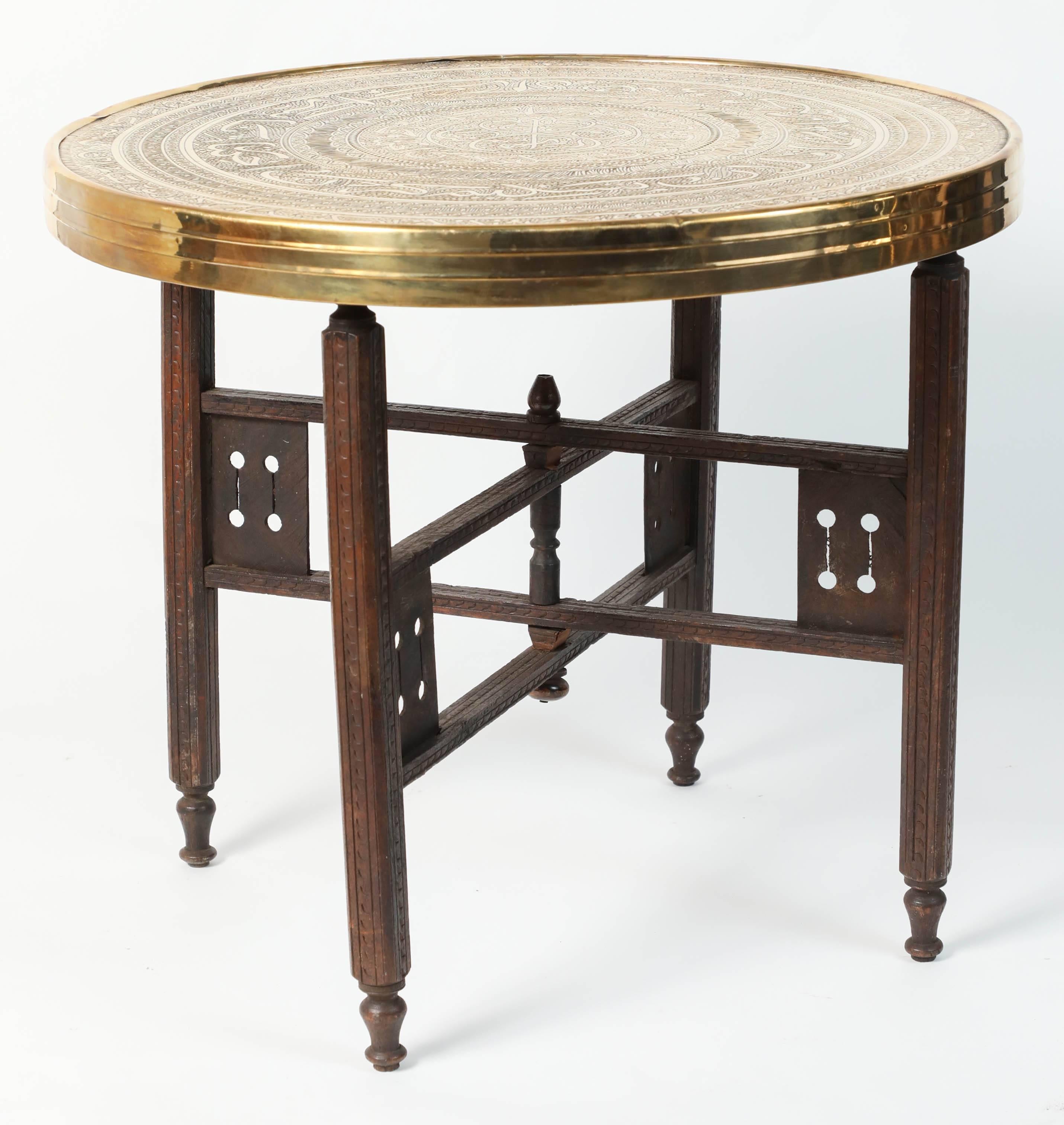 Middle Eastern antique brass tray table with hand-carved wooden folding stand.
The embossed Art Deco style brass top is decorated with Arabic calligraphy writing and repousse geometric Moorish designs.
The top is removable and consist of a round
