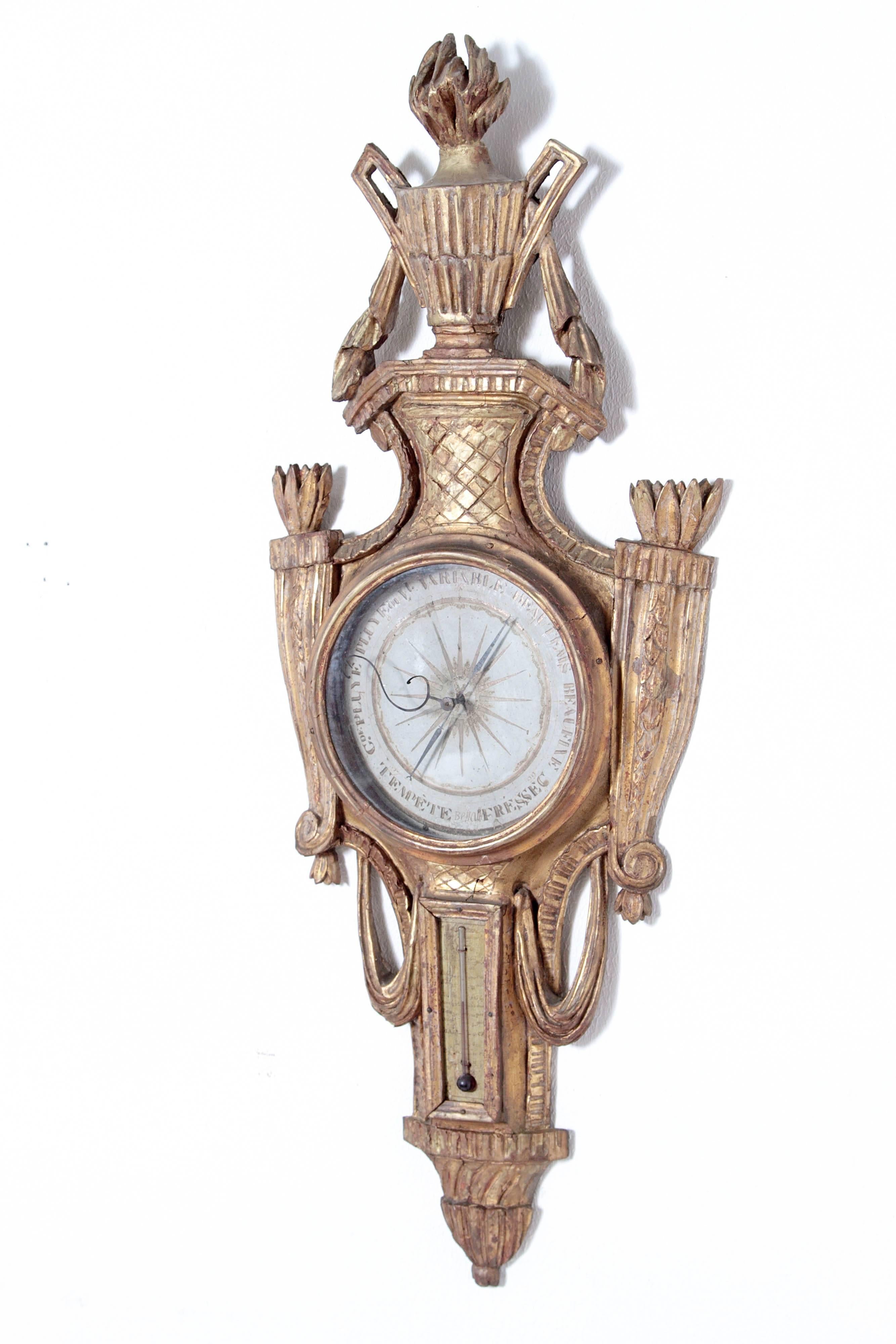 A large French Louis XVI giltwood barometer and thermometer. This piece features a stylized urn with flame at top. The circular face is flanked by carved torches and flames. Functioning spirt / alcohol thermometer below with carved urn form at base.