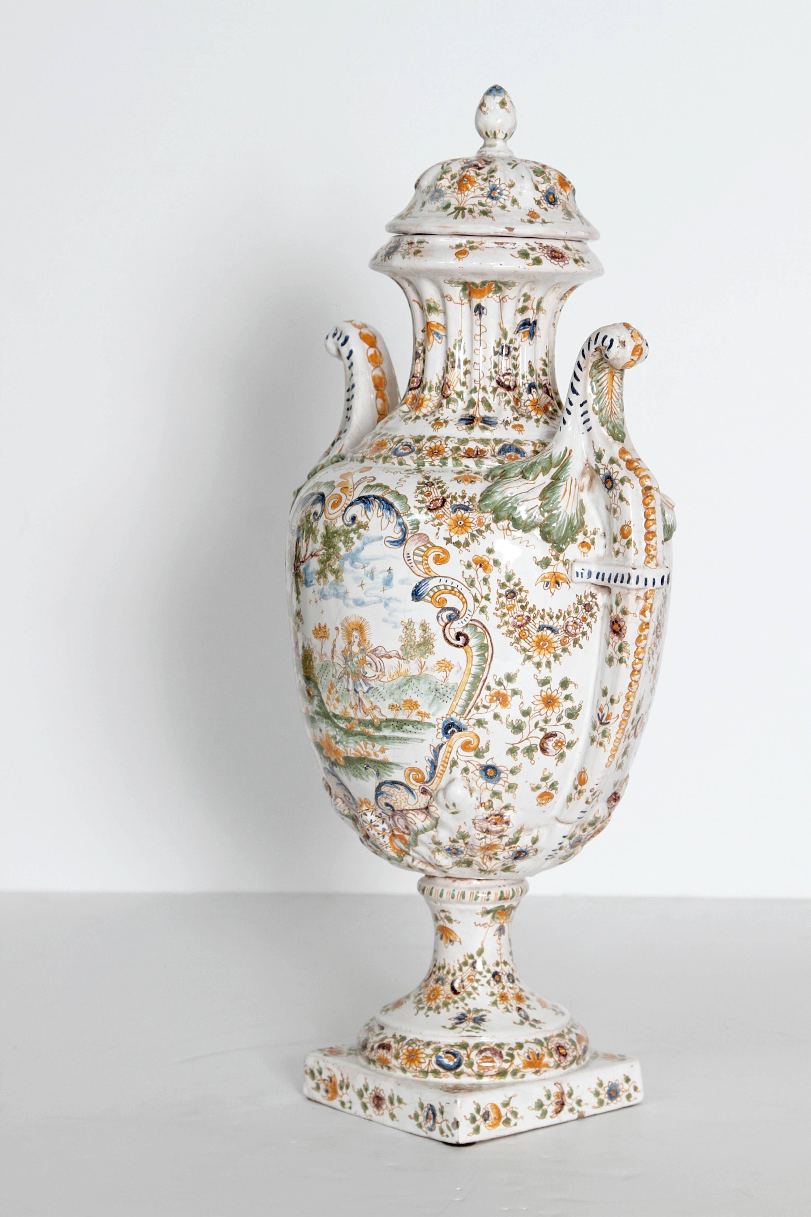Large 18th century French faience lidded urn, white with yellow and blue floral decoration and scenic panels. All this rests upon a 5 inch square pedestal base with floral motif. Curved handles.