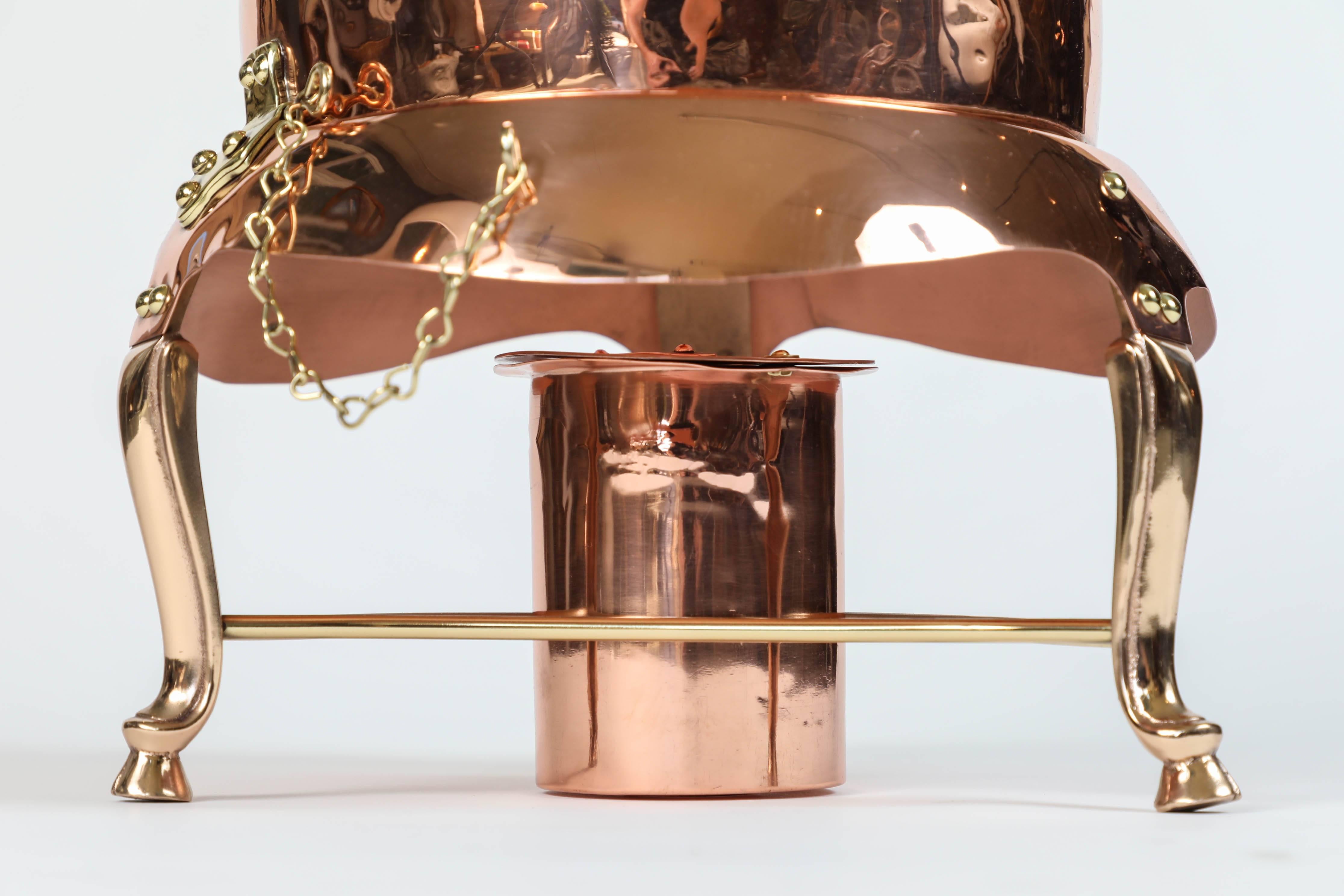 Newly polished vintage large two-gallon copper and brass coffee urn made by J. C. Moore, California Coppersmith. This piece features his trademark hand-carved flame handle. Mounted on brass legs and chained to tilt for easy pouring with matching