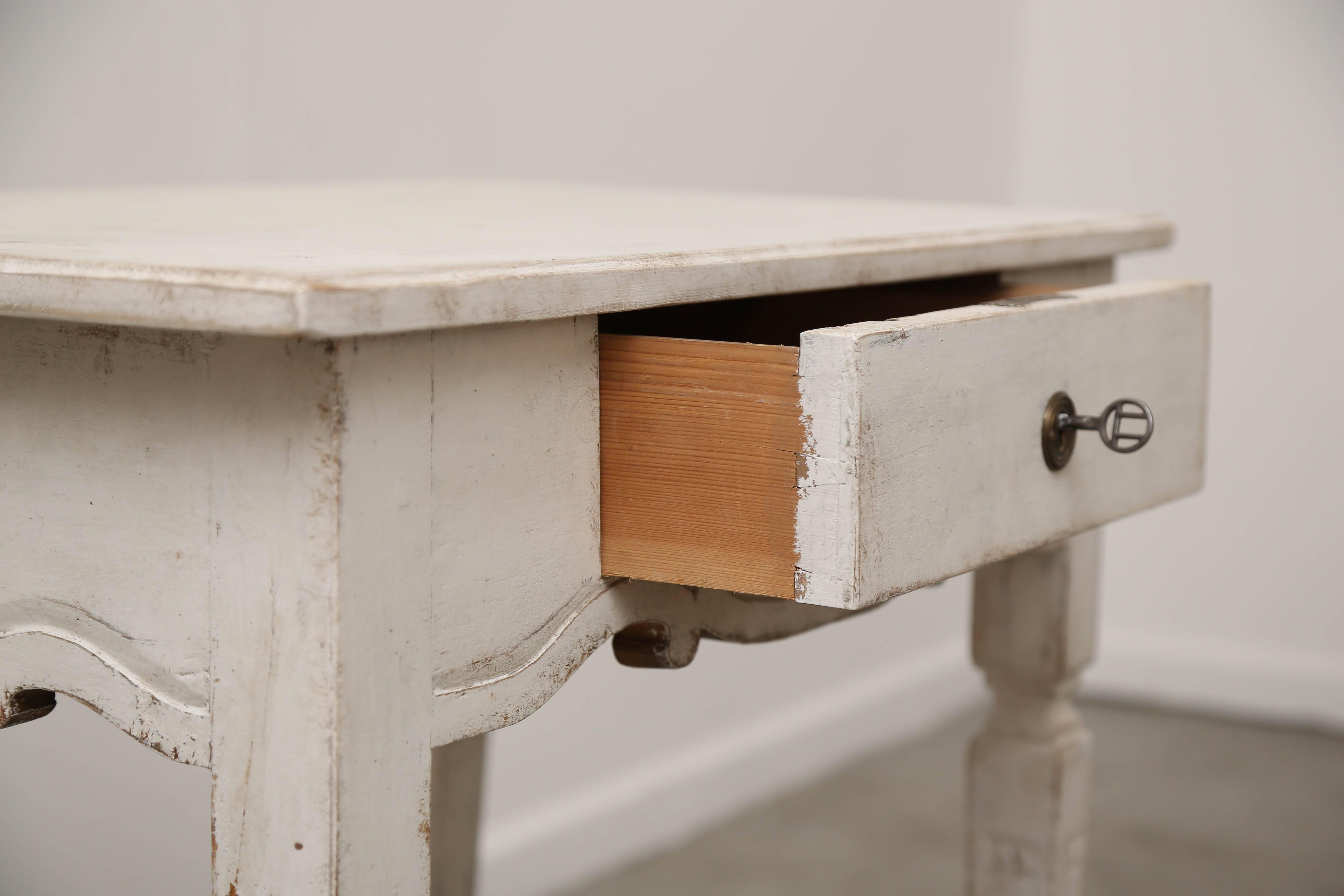 Antique Gustavian style painted side table with single drawer, 19th century, distressed and refreshed Gustavian white/ ivory paint.  That sits on tapered fluted legs with triangular carving at top and the curved apron has turned carved wood