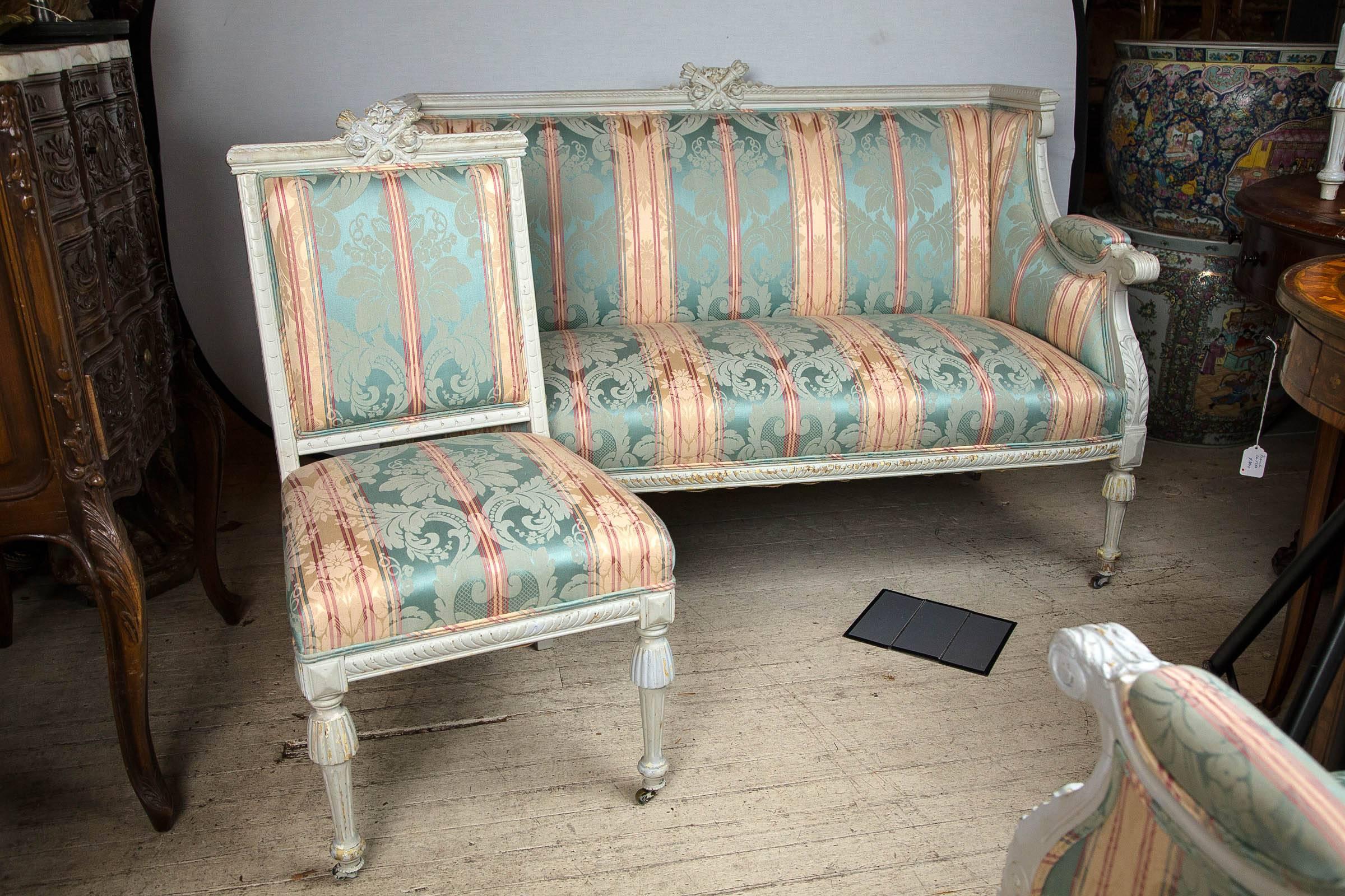 This suite comprises a settee, two armchairs and one side chair. Recently repainted and upholstered. Carved crest on each top rail of crossed torches. Padded arms, acanthus leaf on arm supports. Carved legs and apron. The two armchairs are of