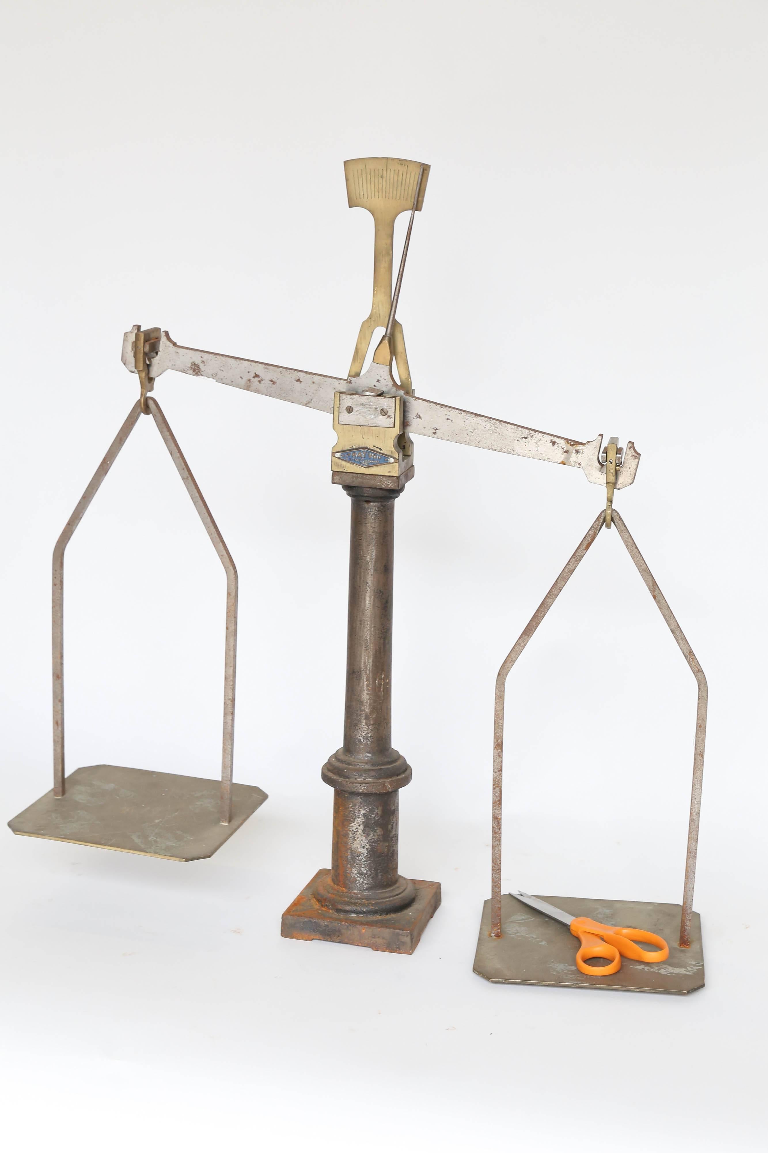 An iron and brass equal-arm scale from the Trayvou Company of Paris and Lyon. The heavy iron pillar supports a steel balance beam with brass fittings and a brass pointer with balance marker. The two brass pans are 7 inches by 7.75 inches. Use it as