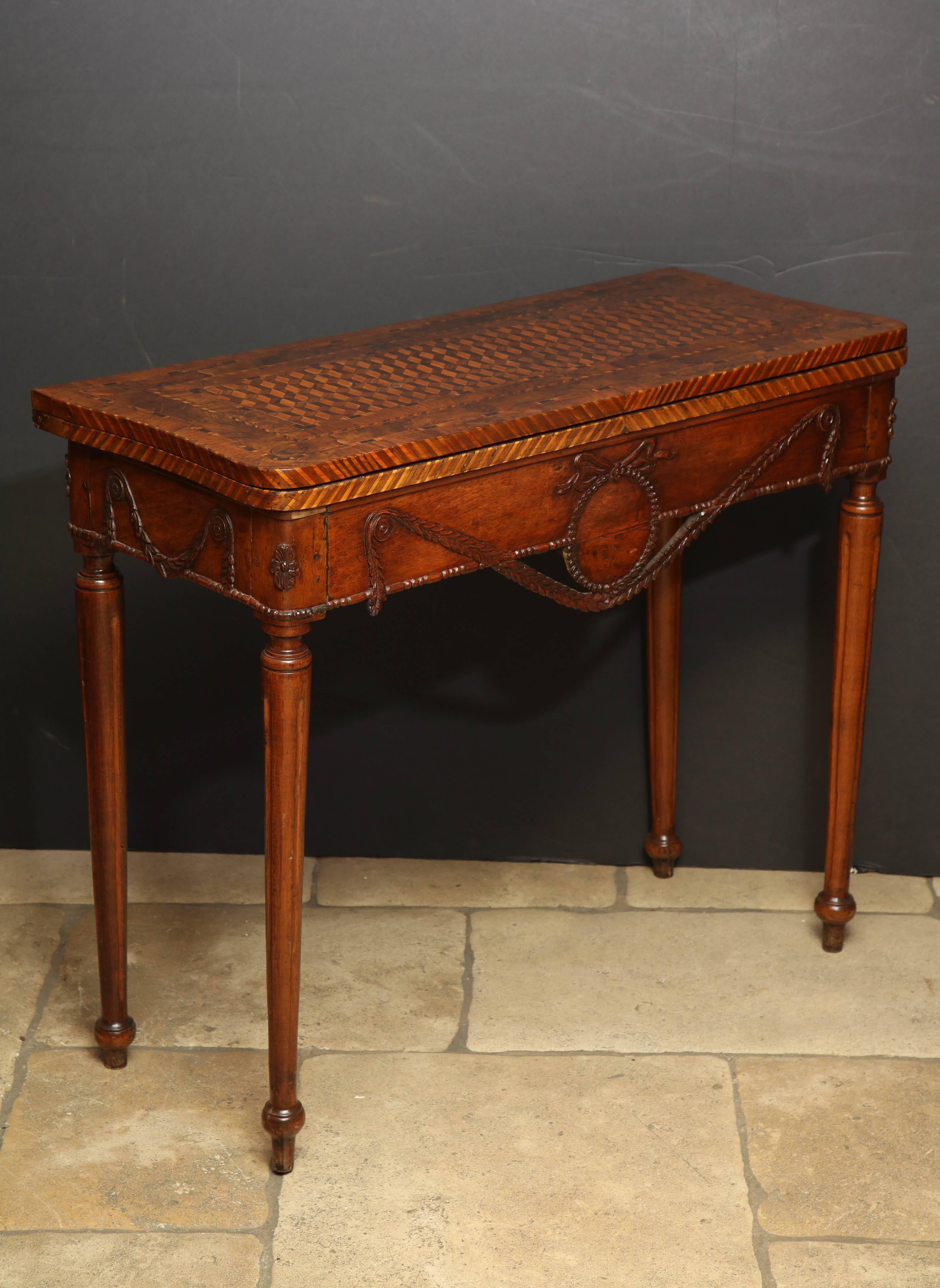 An unusual Louis XVI carved walnut flip-top game table with parquetry inlaid veneers, carved bownkot and garlands about turned fluted legs.