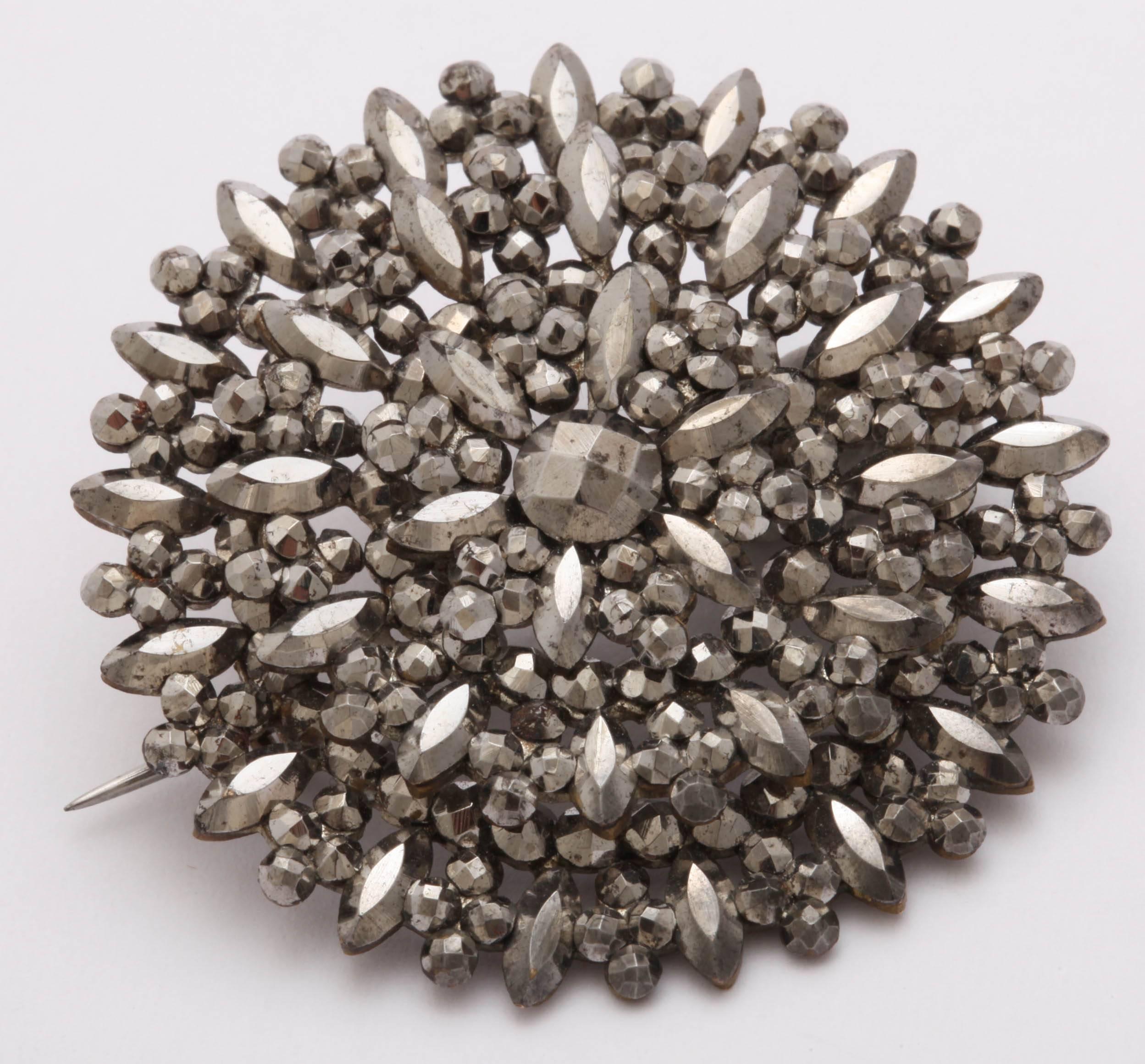 Der geschliffene Stahl dieser Brosche besticht durch die Vielzahl der Formen, aus denen sich dieses stark strukturierte Design zusammensetzt. Die Mitte ist wie ein Diamant im Rosenschliff geschliffen. Ringsherum sind polierte Stahlkugeln und Kugeln