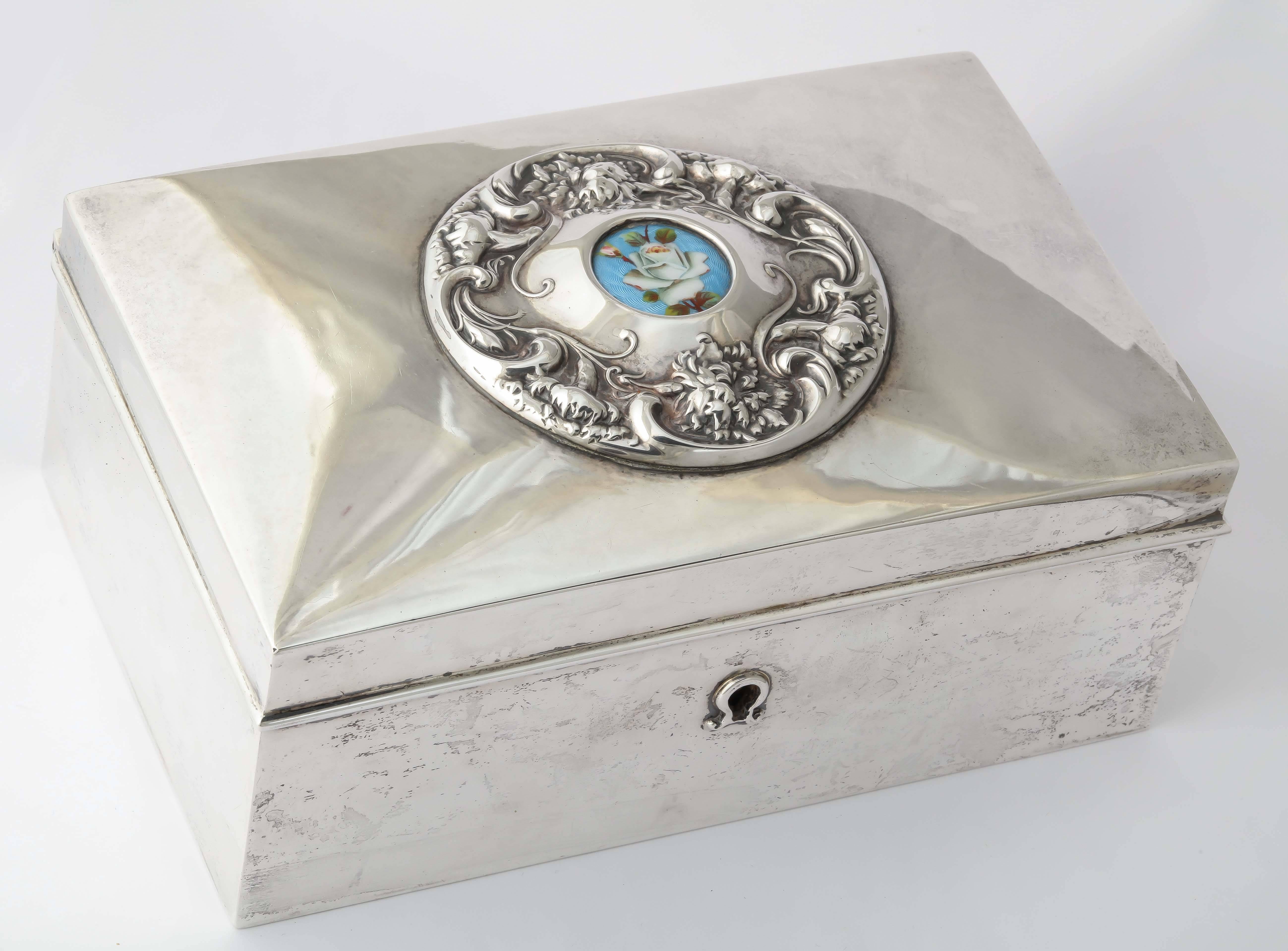 Ein schöner rechteckiger Liebesbriefkasten aus schwerem Silber mit einem Schlüsselloch. Der Deckel ist mit einem kreisförmigen, erhabenen Blattmotiv verziert, das eine emaillierte weiße Rose auf blauem, guillochiertem Grund einschließt. 

Von