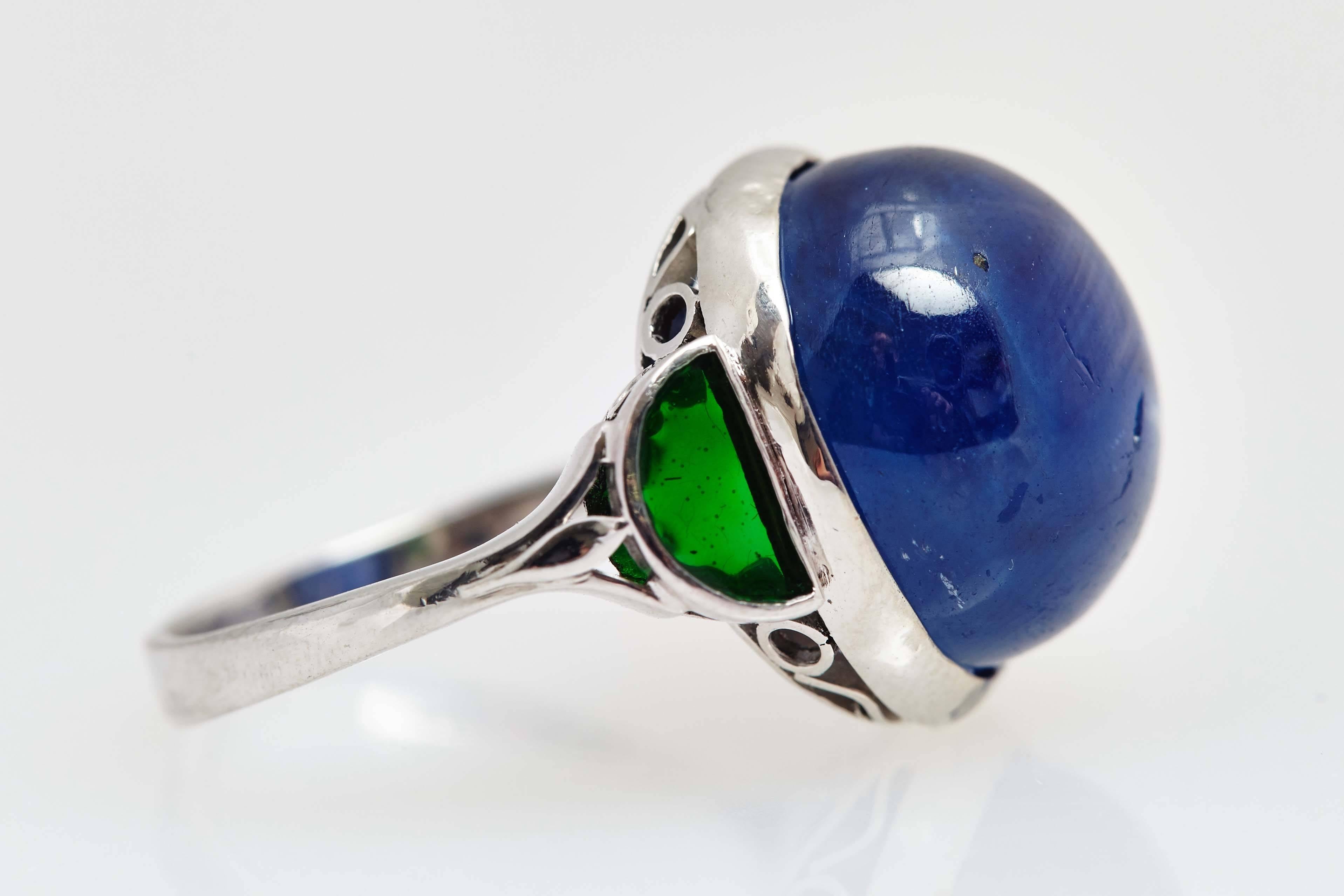 A platinum ring showcasing a cabochon Burmese blue sapphire - non heated- weighing 18.91 cts, sided by two half moon shaped emeralds. Circa 1965 