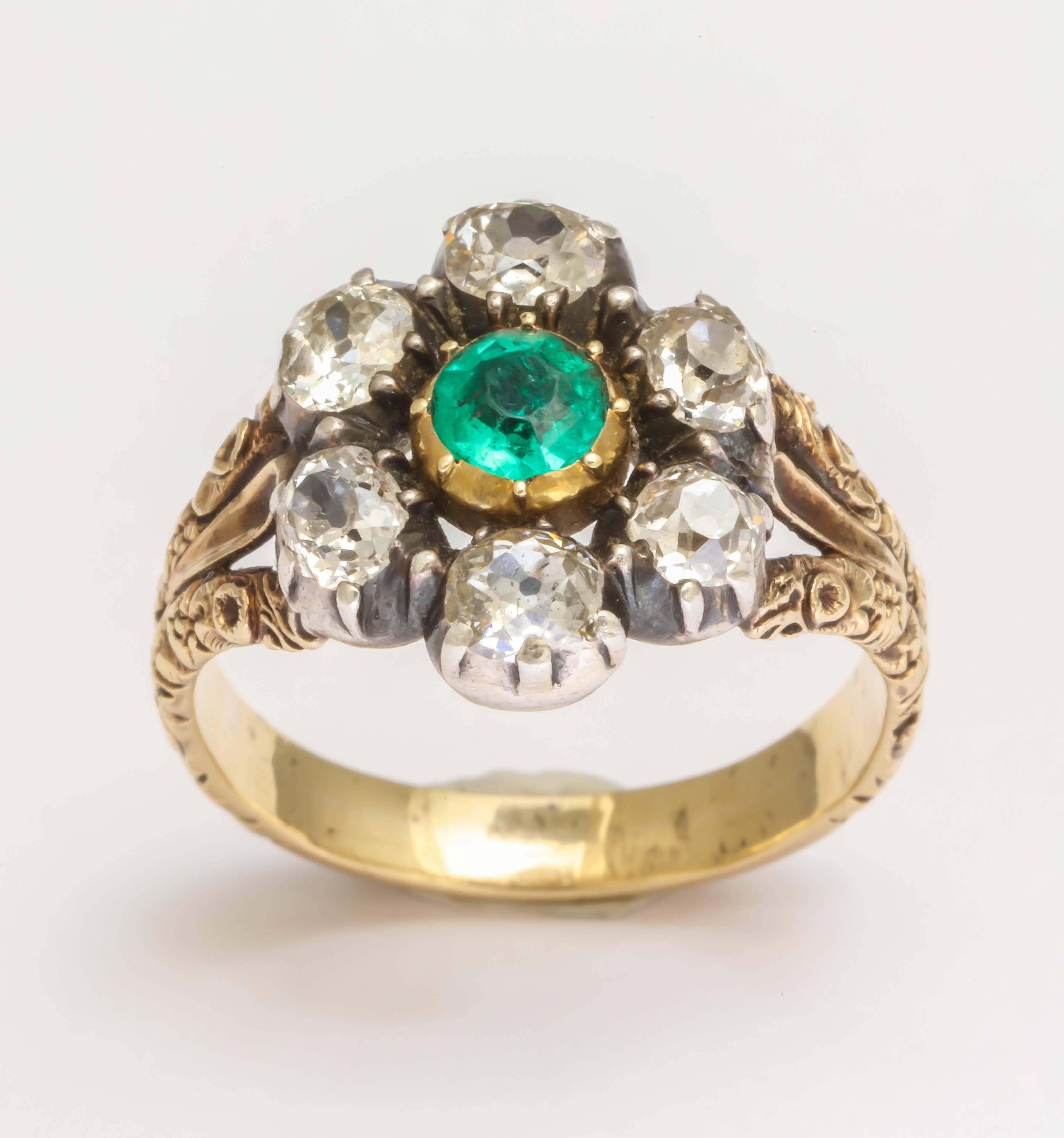Gorgeous emerald is surrounded by six old mine cut diamonds in a cluster closed back setting. The mount has lovely split shoulders with engraving throughout. Silver over yellow gold, likely made in England. 