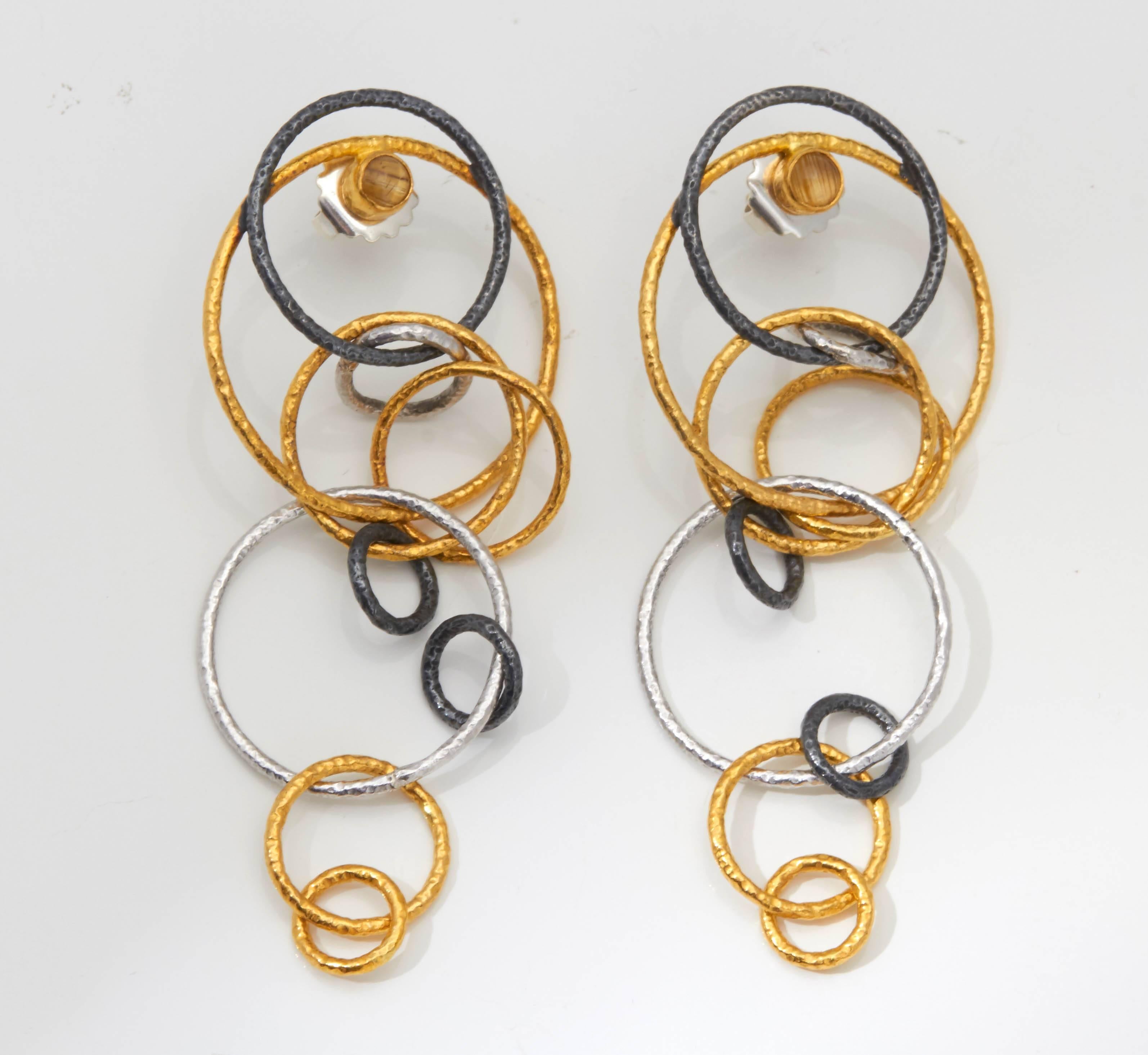 A pair of earrings composed of varying sizes of rings in 18kt yellow gold, sterling silver and rhodium plated silver. There is a bezel set chrysoberyl at the top of the earring.
Length: 3.50 inches
Width: 1.40 inches