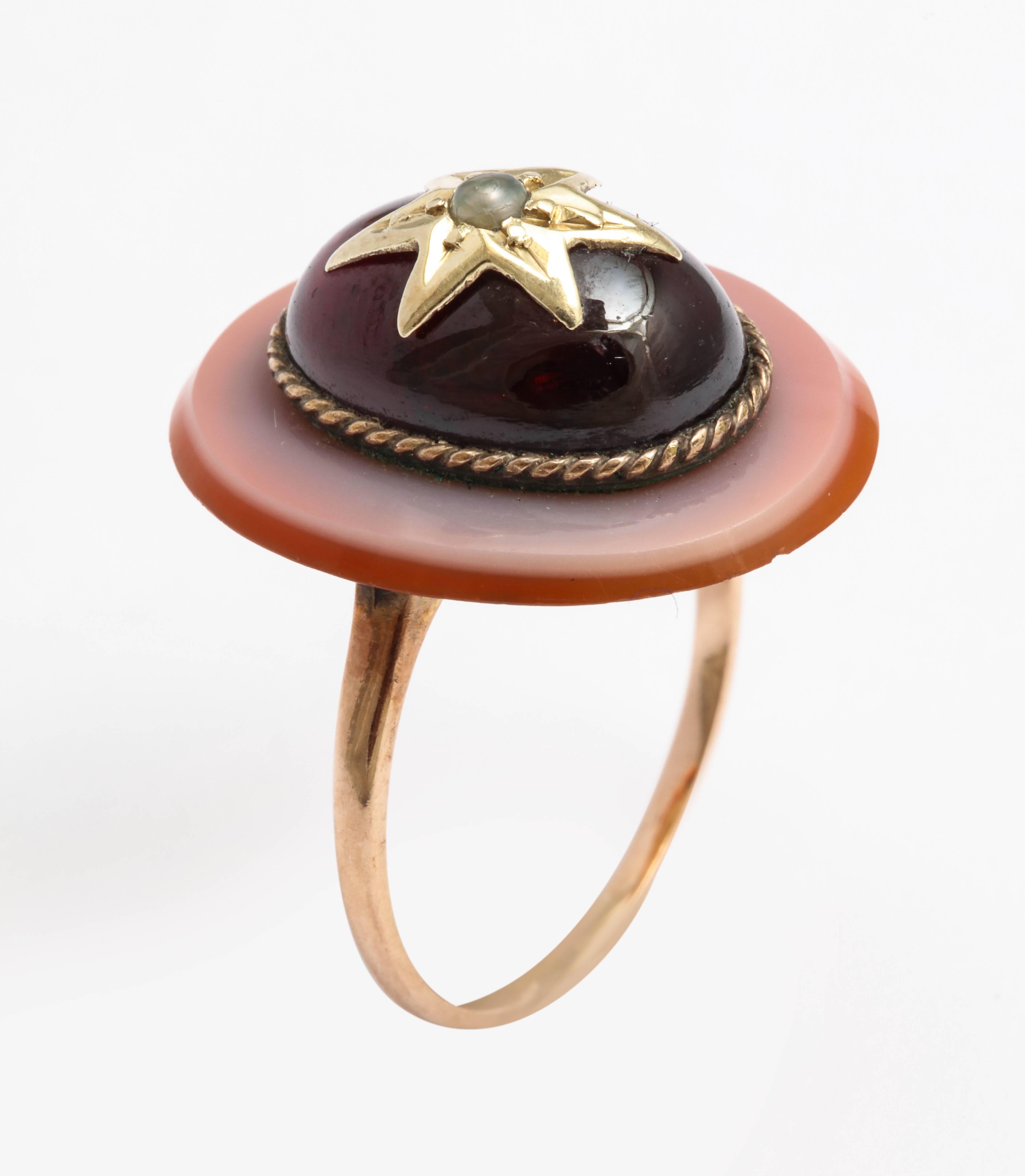 Set with a cabochon garnet, the center with a gold star set with a natural seed pearl, mounted on an oval banded agate base, separated by a twisted ropework band, the shank of 10k gold. 

Circa 1890.

size 5. 

Garnet measures 14.2 x 10mm; agate