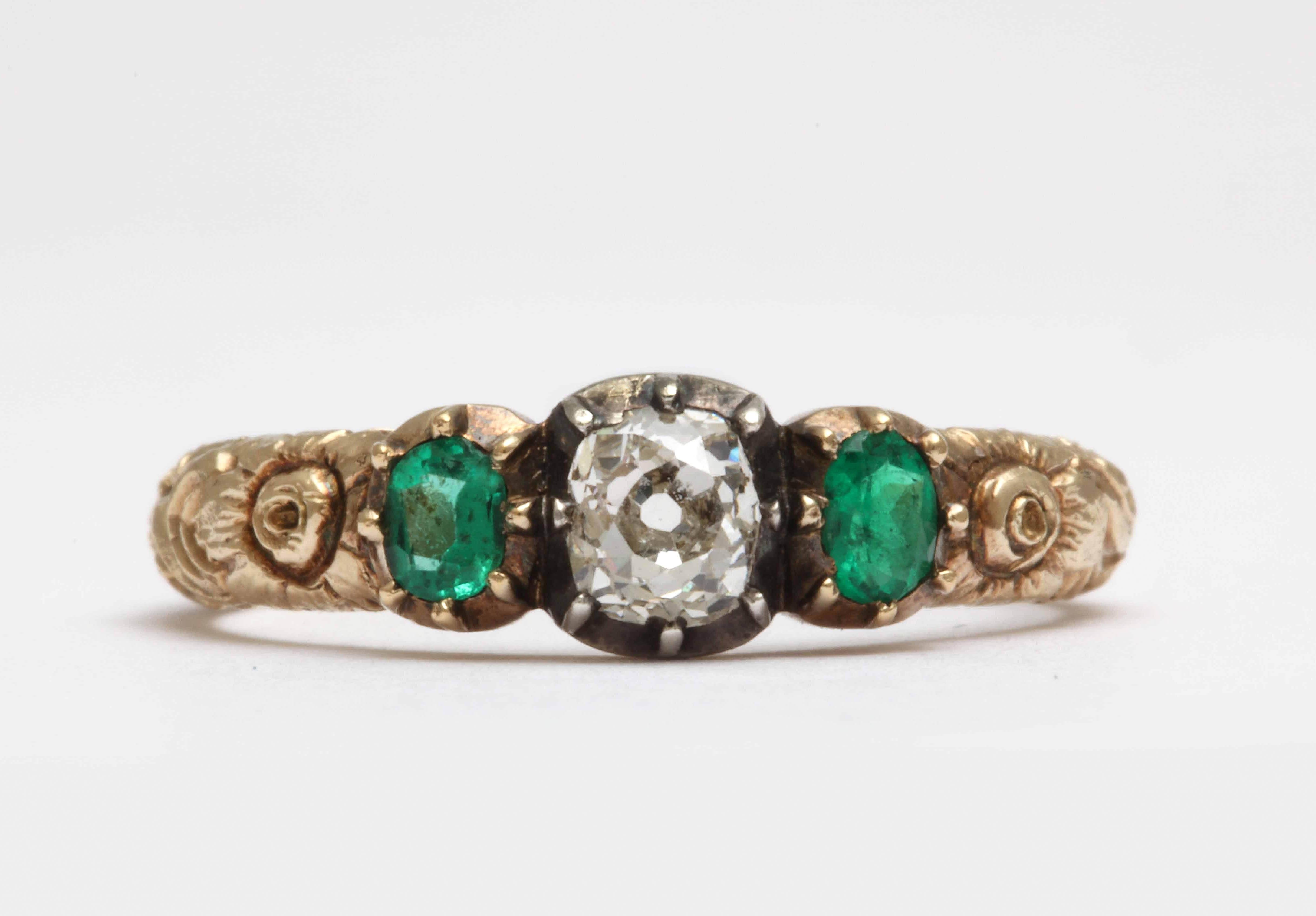 Made in the style of Georgian rings, but the stones are set with open backs. A central old mine cut diamonds is set in silver flanked by two emeralds set in yellow gold. The shank has beautiful deep floral engraving all the way around. 15kt-18kt
