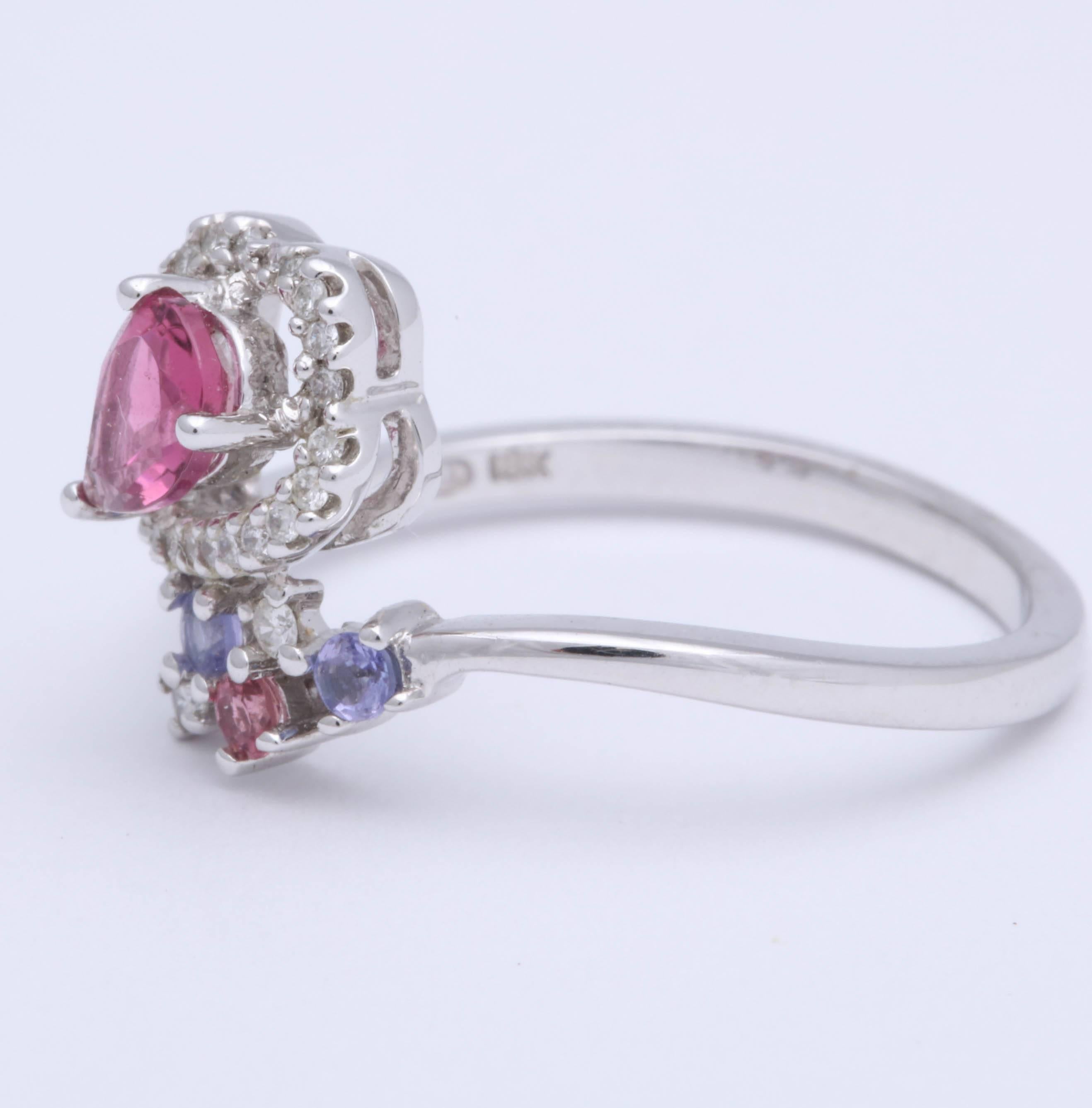 Sweet and stylish pear shape pink tourmaline ring in 18 kt white gold. The tourmaline is enhanced by periwinkle colored round tanzanite and a pink sapphire. A row of diamonds surrounds the center stone, total diamond weight is about .26 cts. The