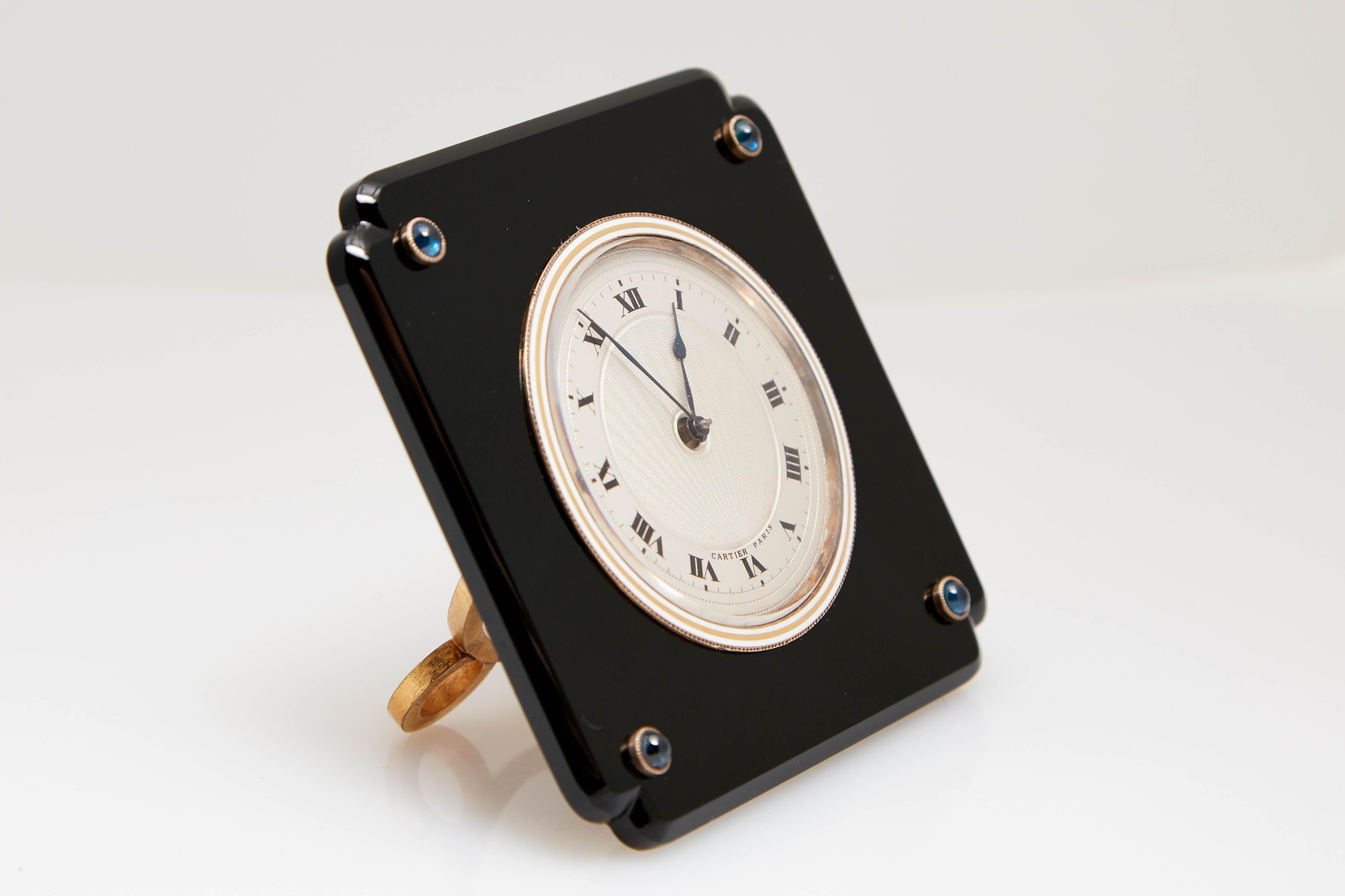 Cartier Art Deco black onyx and cabochon sapphire desk clock. Made in Paris, circa 1925. Mechanical, the circular matte silver-tone dial centering an inner circle of engine-turned sun ray pattern, with black Roman numerals, blued steel spade hands,