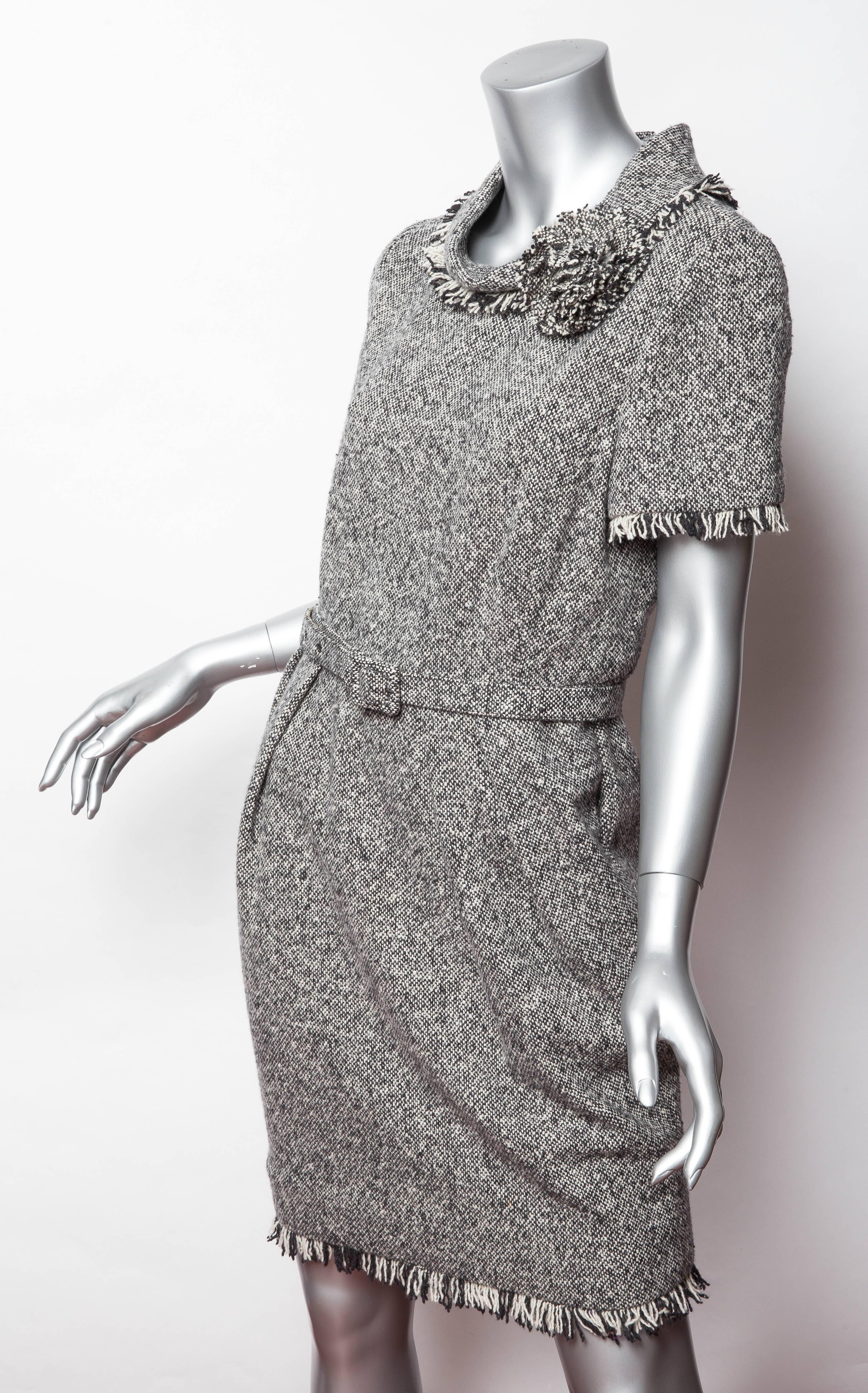 Very chic tweed short sleeved dress with cowl neck and corsage detail. Sleeves, hem and neck are trimmed with fringe. The bottom of the dress is slightly ruched to one side and an adjustable belt in the same fabric as the dress serves to highlight a