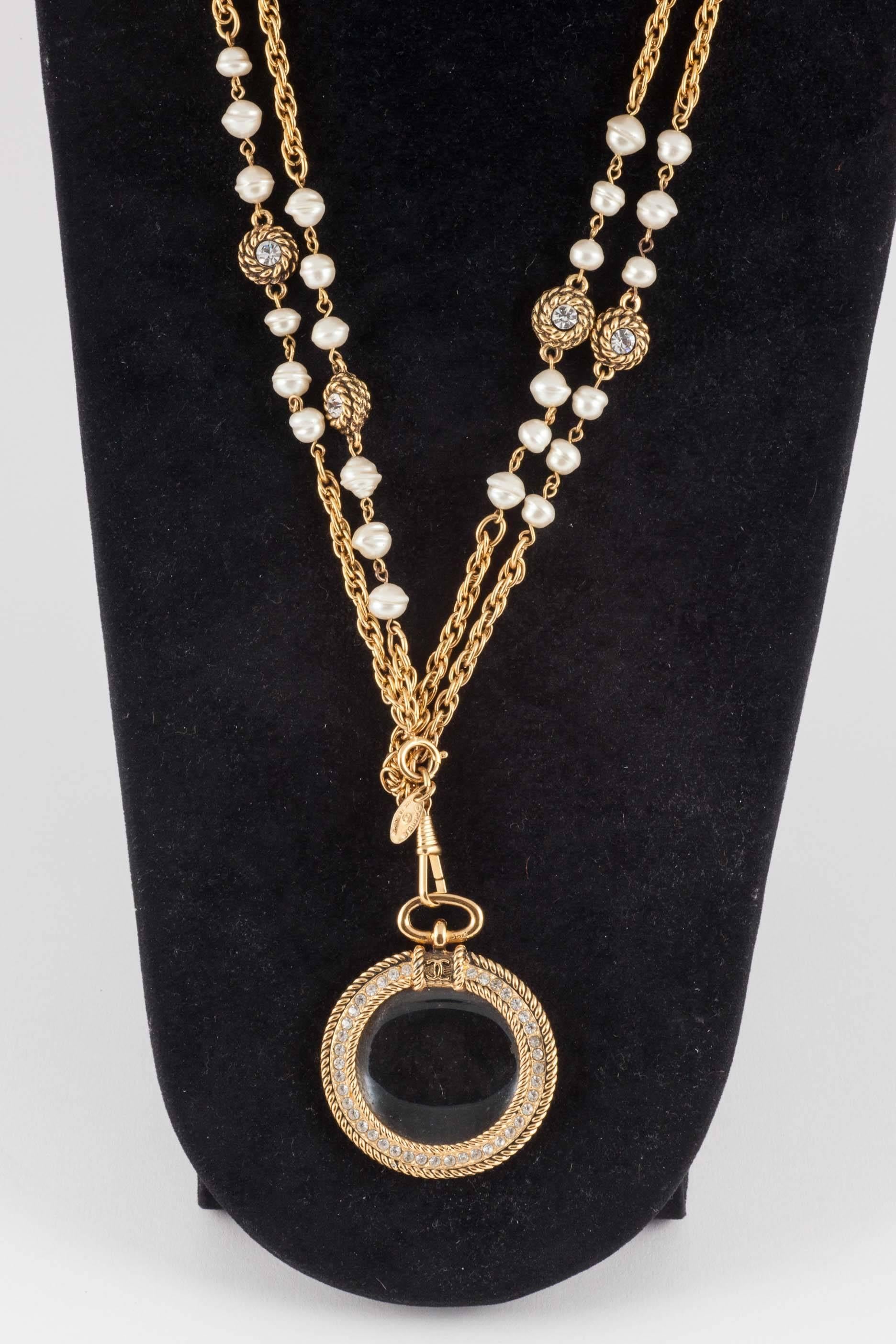 Quirky and chic pendant necklace from Chanel, in the 1980s. Highly characteristic of this period, it is made from glass baroque pearls, paste and gilt metal chain, sporting a most useful magnifying pendant. This highly popular and iconic necklace