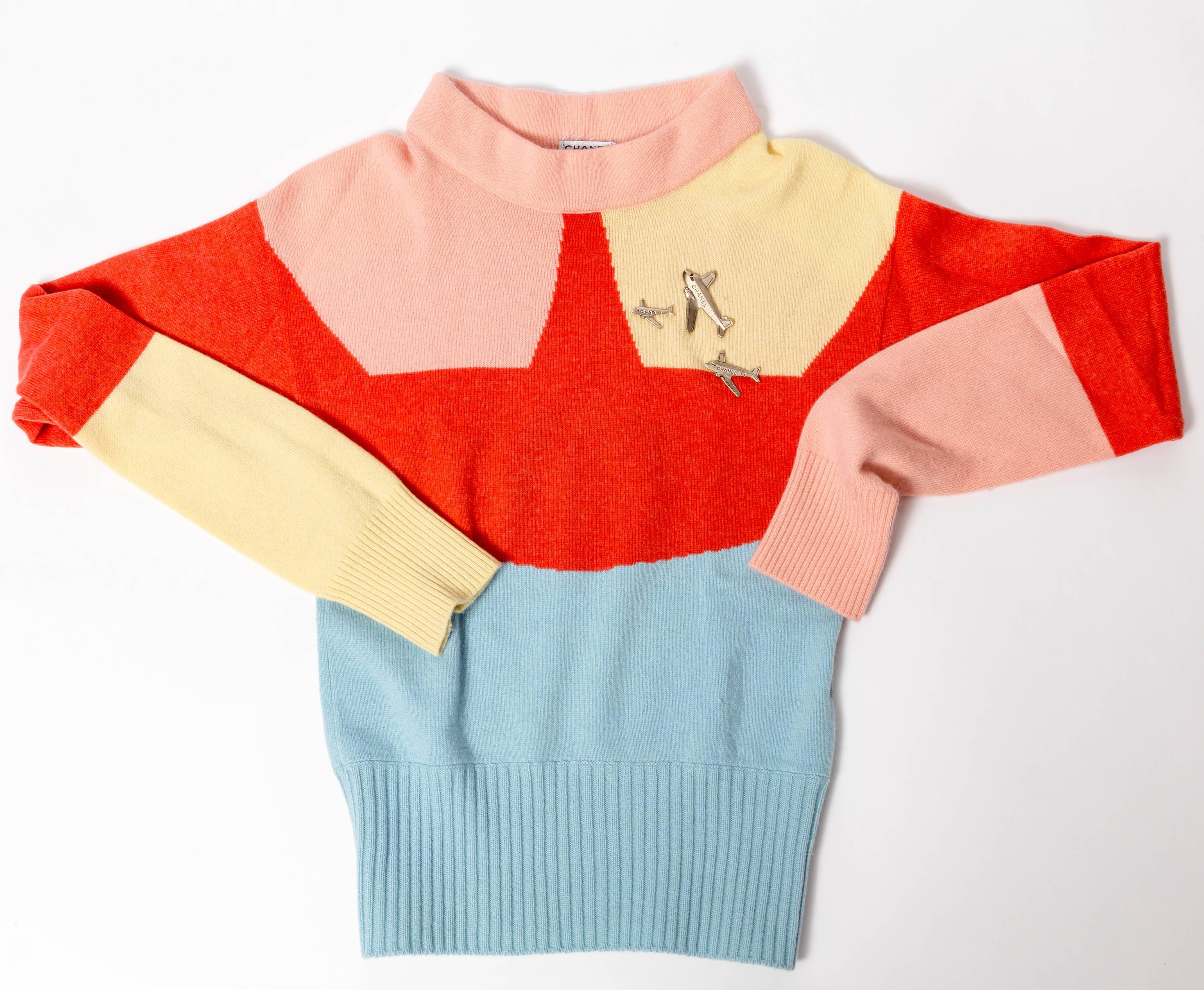 Very chic Chanel cashmere color blocked sweater with airplane charms.
In shades of blue, pink, orange and yellow with a charming cowl neck collar. P32421K00694
Airplane charms in silver metal and signed CHANEL are sewn on to the left breast of the
