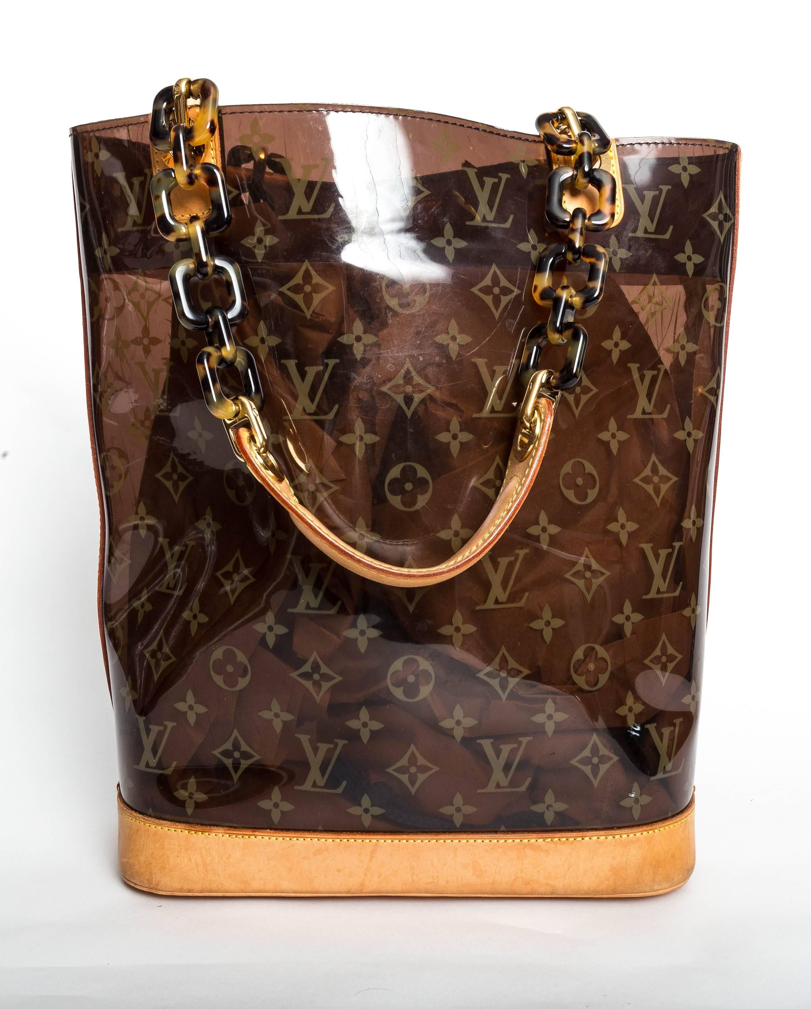 This chic tote is crafted of Louis Vuitton monogram on amber transparent vinyl. The bag features tortoise chain top handles with a vachetta leather grip, brass links, leather anchors, and a full vachetta leather base. The top is open to a spacious