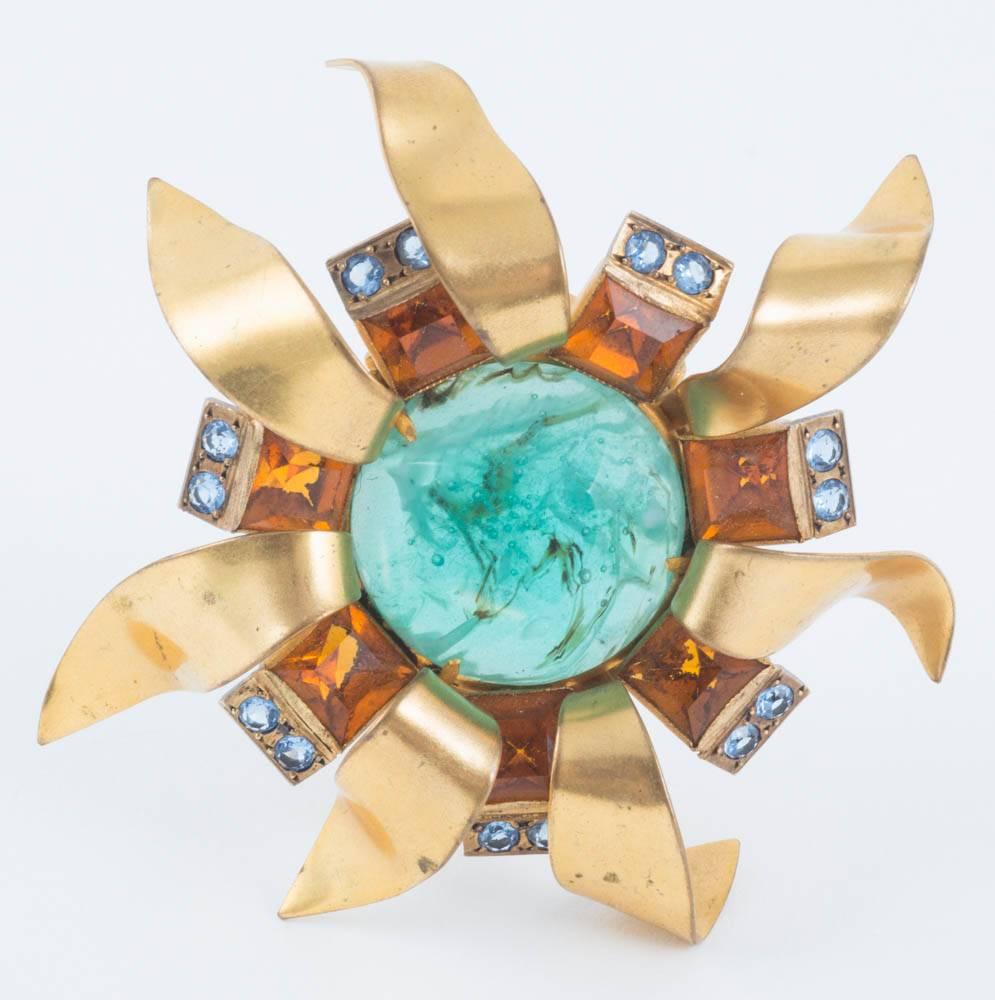 A fabulous hand crafted brooch depicting a stylised sunburst, or sunflower, with a large resin 'emerald' centre stone (with inclusions!), surrounded by square cut topaz pastes and light blue round pastes. The 'leaves' or 'sunbursts' are individually
