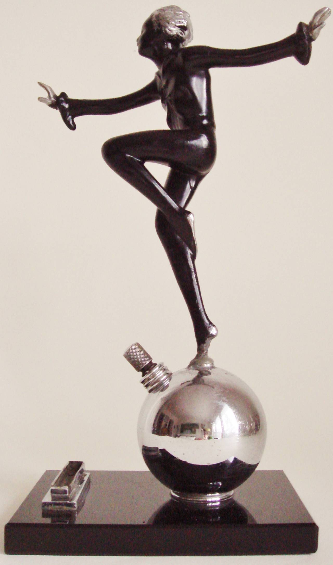 This rare English Art Deco figural striker lighter features a cold-painted Joseph Lorenzl black and chrome female figure who is balancing with arms outstretched on a chrome sphere that houses the striker wand.This sphere along with the striker strip