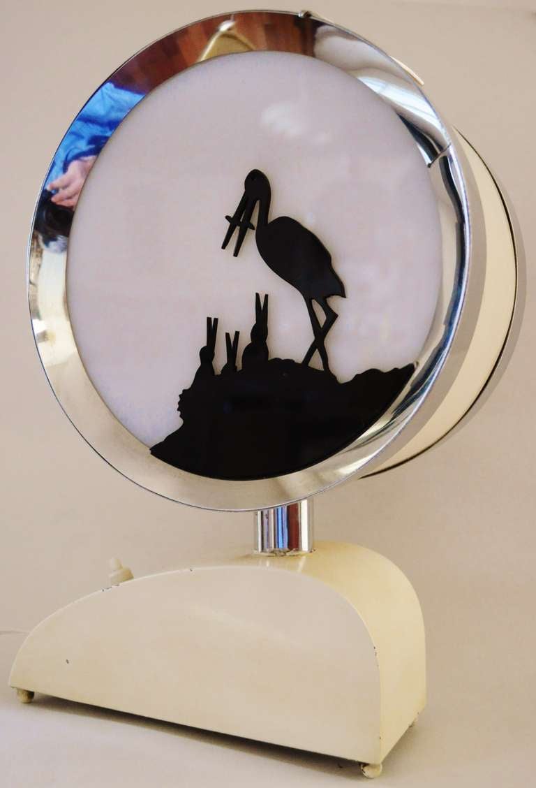 This amazing English Art Deco nursery night light or table lamp is in the shape of a chrome banded white enamelled bass drum. One face of the drum features a black Lucite silhouette of a stork on the nest feeding her young, while the other face