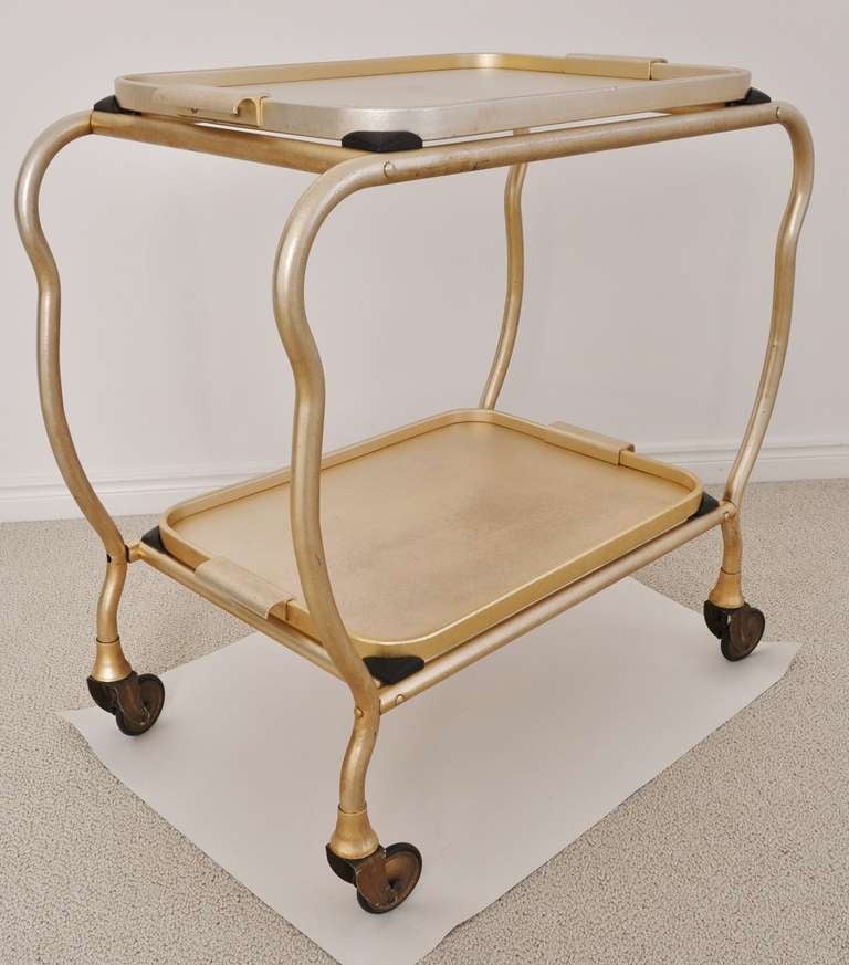 This is an example of the scarce “cloud profile” English cocktail cart or tea trolley in anodized gold. It has two large detachable trays and independent patented “Flexello” sprung and rubber tired wheels, that enable the cart's owner to serve tea