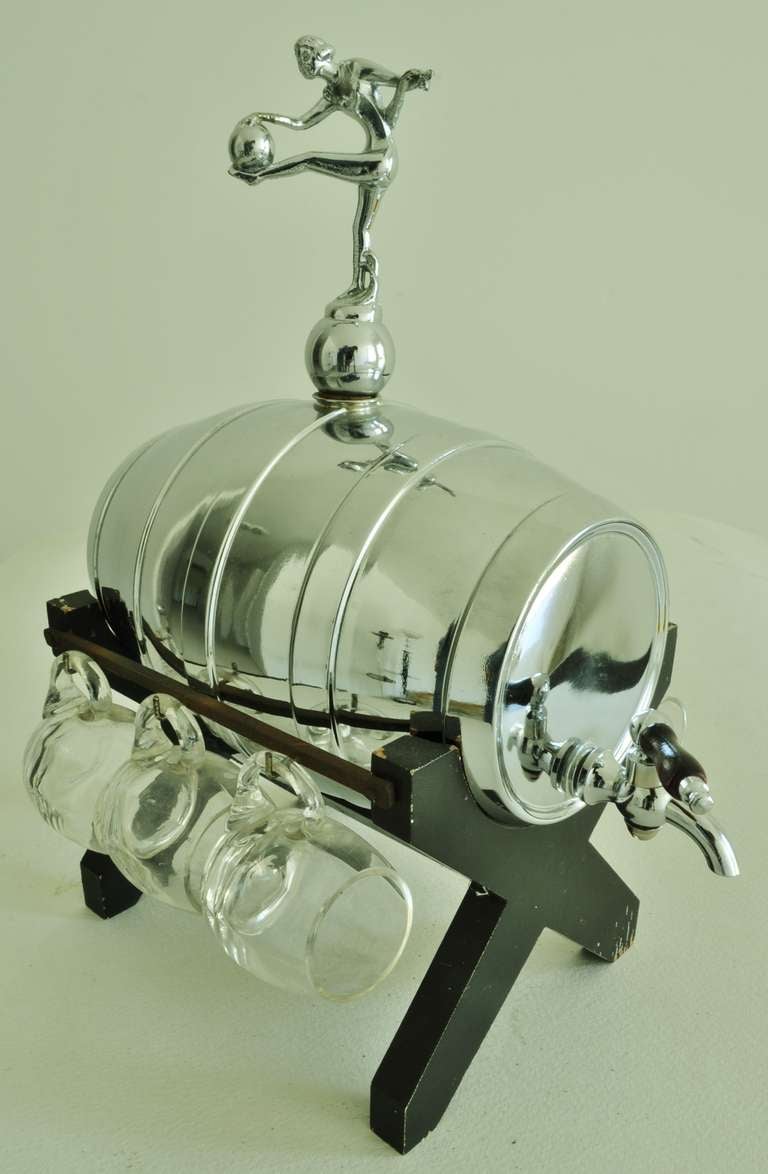 This wonderful English Art Deco chrome plated barrel shaped whisky dispenser is complete with its six original miniature beer mug shaped glasses. The spigot and cross-braced stand are lacquered wood and the cork “bung” at the top sports a nude