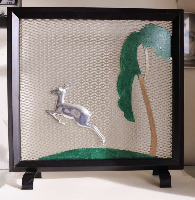 This wonderful Art Deco fire screen features a leaping gazelle in high polished aluminium on an aluminium mesh background featuring a green hillock with a brown and green tree. The frame, legs and handle at the back are all stove enameled in black.
