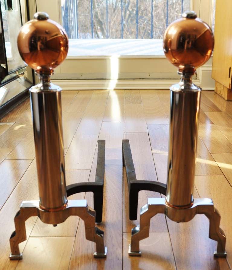 This pair of beautifully restored, iconic American Art Deco chrome, copper and iron fireplace accessories feature a chrome button that attaches a copper sphere to a chrome tubular shaft that terminates in a copper ring before splitting into two