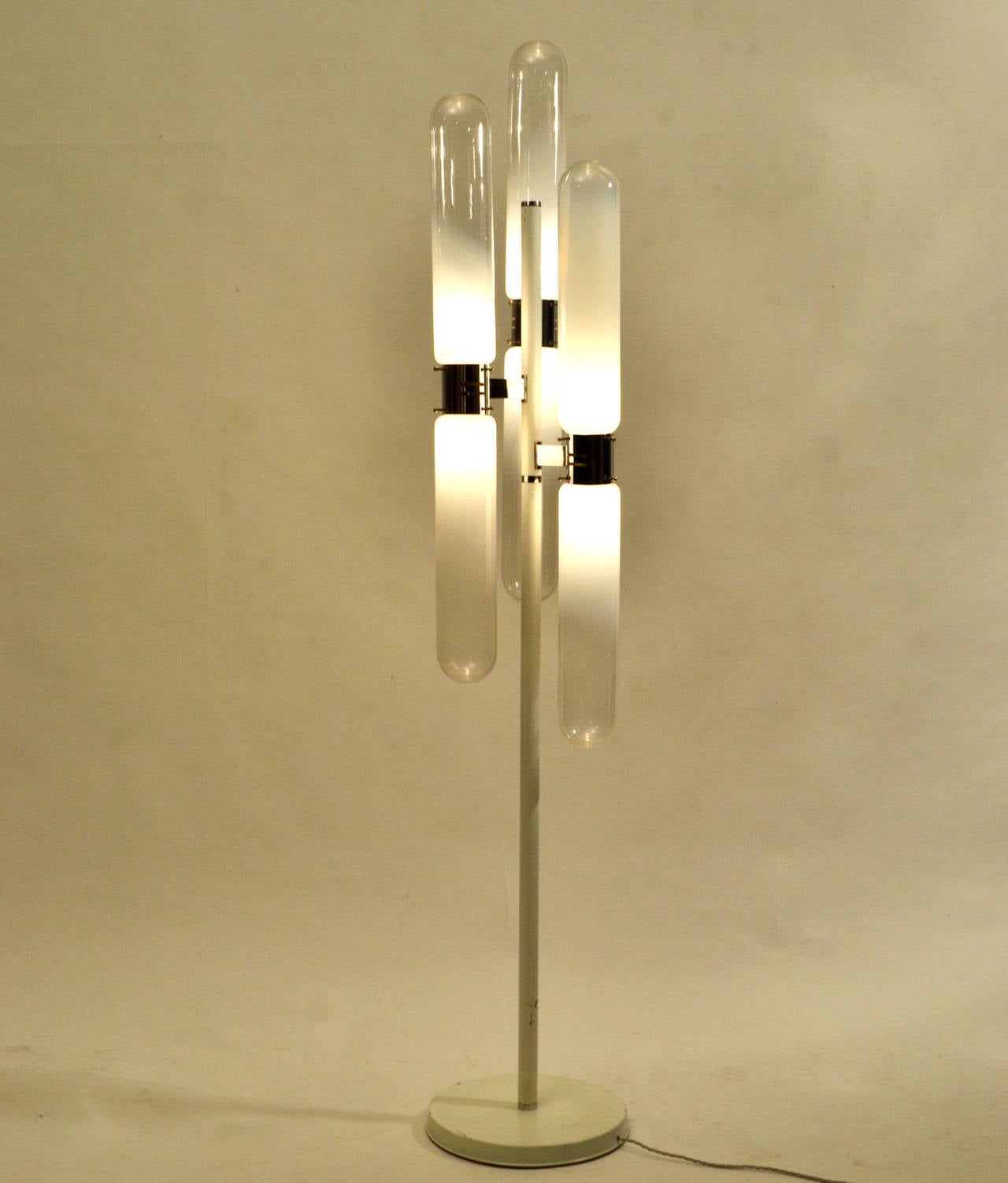 Lampadaire avec six abat-jour en tube soufflé à la bouche, reliés de haut en bas à la structure centrale en métal chromé et blanc.
La couleur du verre va du blanc laiteux au translucide, puis au clair qui diffuse la lumière.