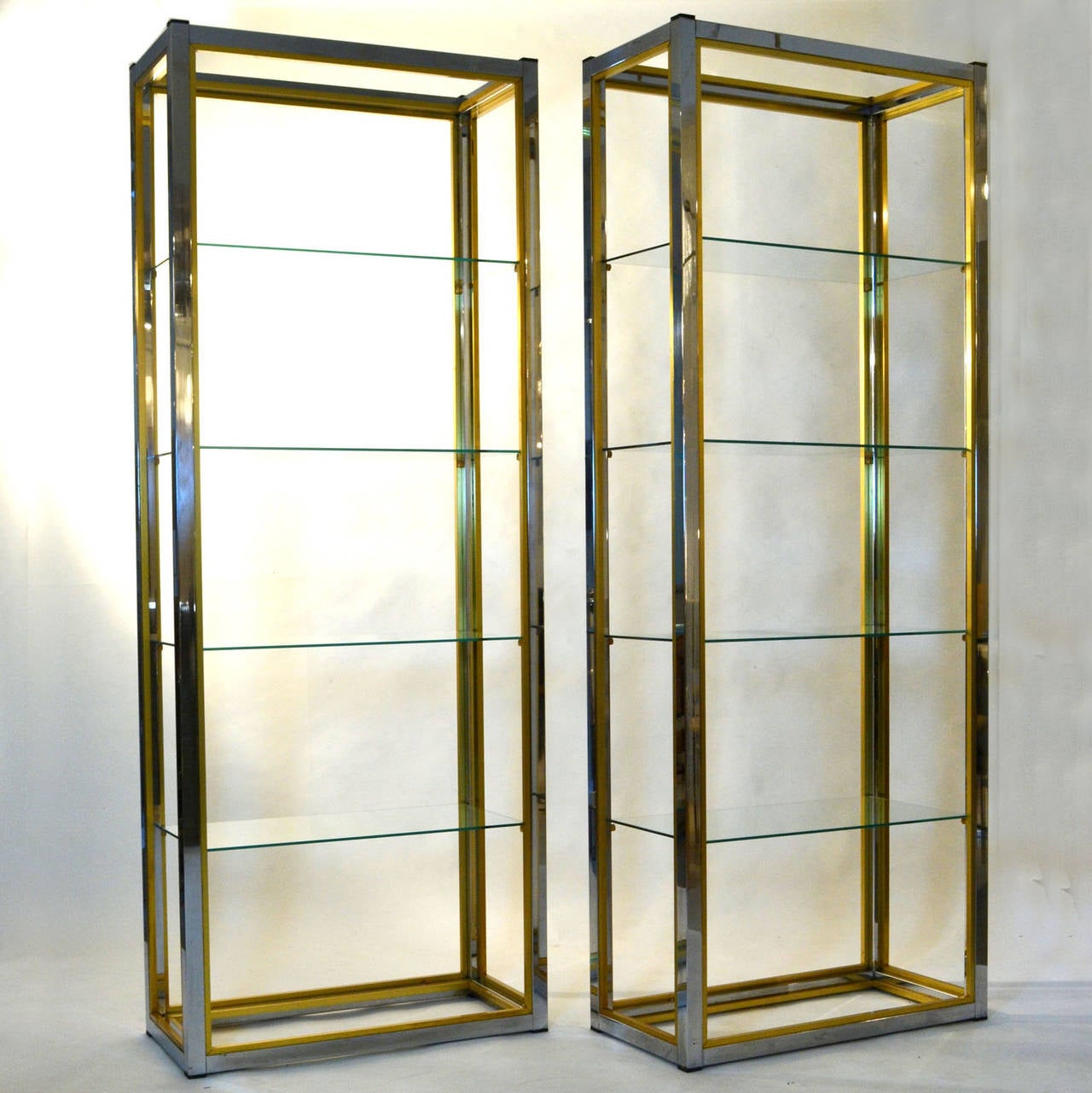 Two rectangular freestanding minimal shelving units are designed with a chrome outer frame and a brass inner frame. They have clear glass shelves.