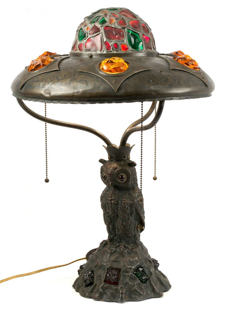 An Art Nouveau bronze and slag glass table lamp with and owl standing at it center. The shade and base are inlaid with colored cut glass in greens, reds, and yellows.