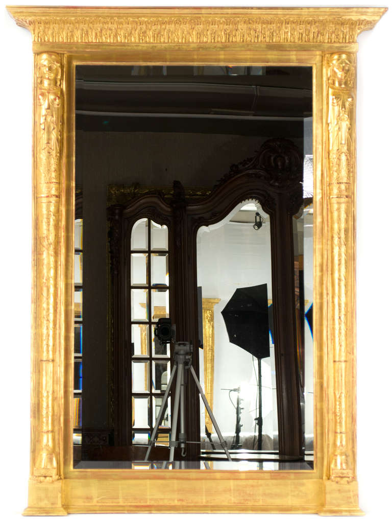 A large gilt and bevelled mirror framed on both sides with Egyptian-inspired caryatids and topped with pediment festooned with dental work and acanthus leaves.