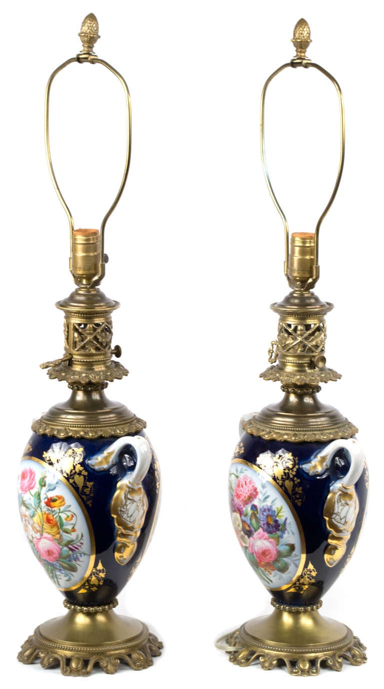 These two vases feature exquisite painted floral cartouches surrounded by dark blue and gold. The vases date to the 1870s and were later (c. 1900)  mounted with bronze bases, lids, and harps. The pair is wired and in fine working condition.