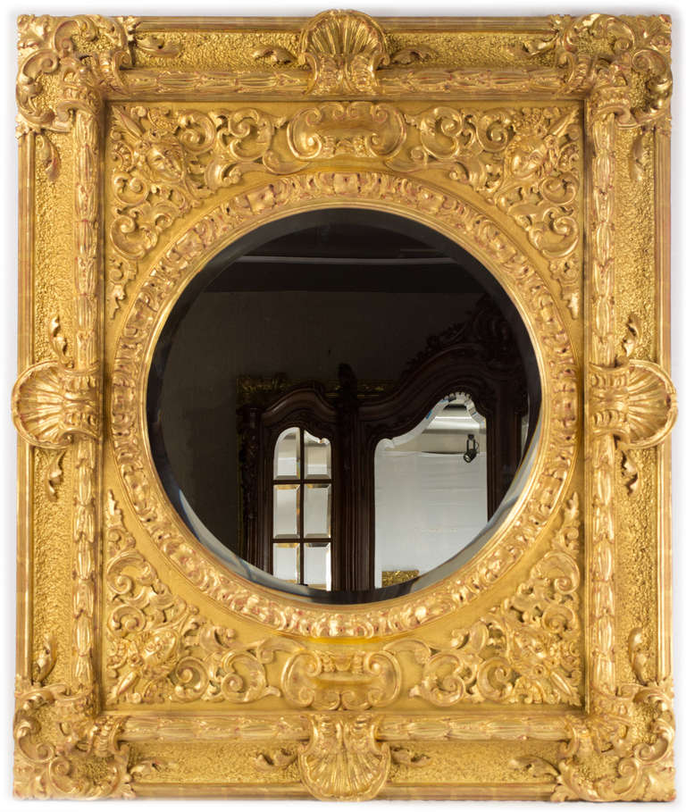 This large, gilt and bevelled mirror is carved with masks, heavy shells, acanthus leaves, c-scrolls, and foliage sprays.